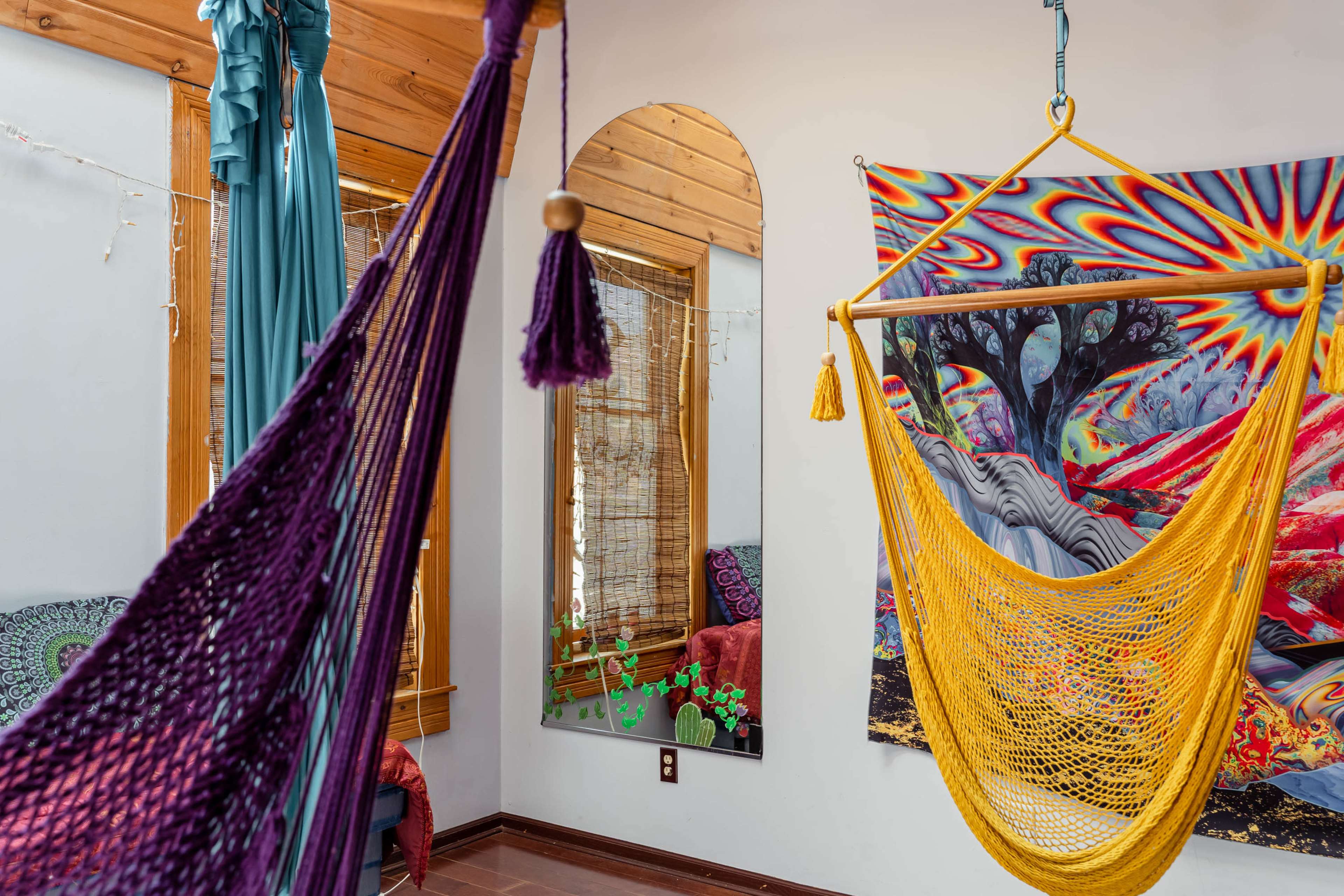 A room features colorful hammocks, a decorative mirror, and a vibrant tapestry against a neutral wall.