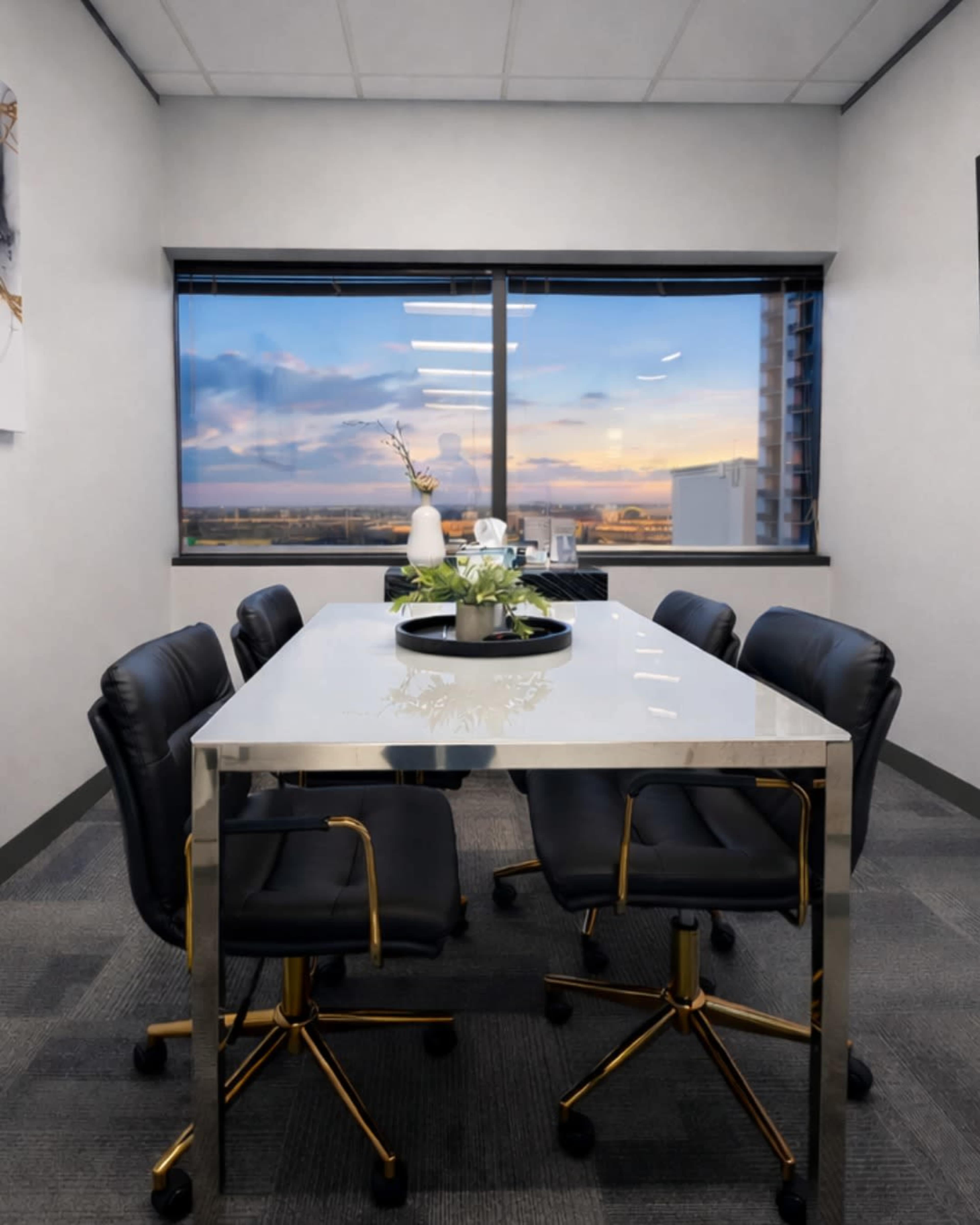 A sleek conference table with black rolling chairs is positioned in a modern office space featuring a large window that offers a view of a sunset sky.