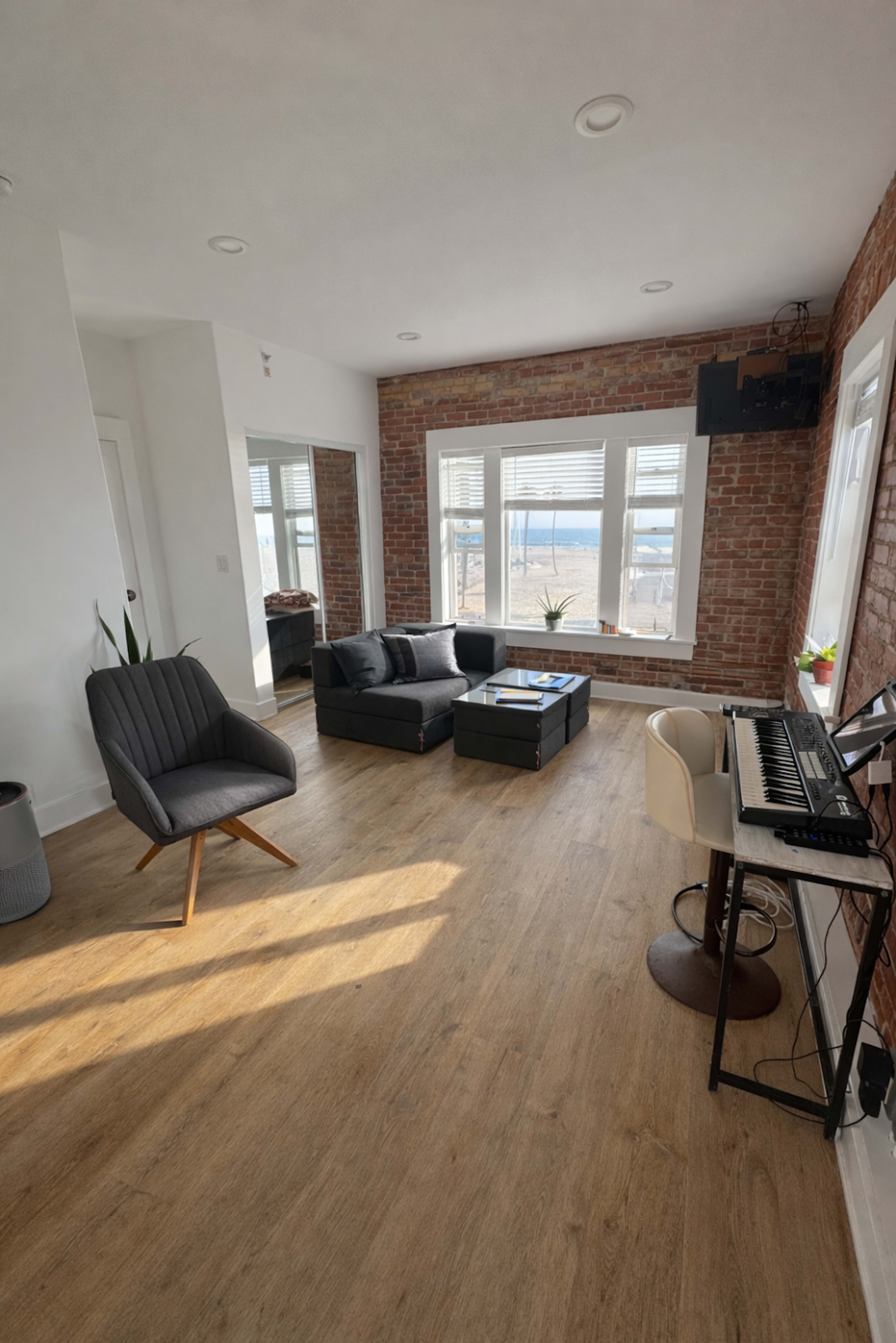 The room features a gray sofa and chair arranged around a coffee table, with large windows offering a view and a piano set against a brick wall.