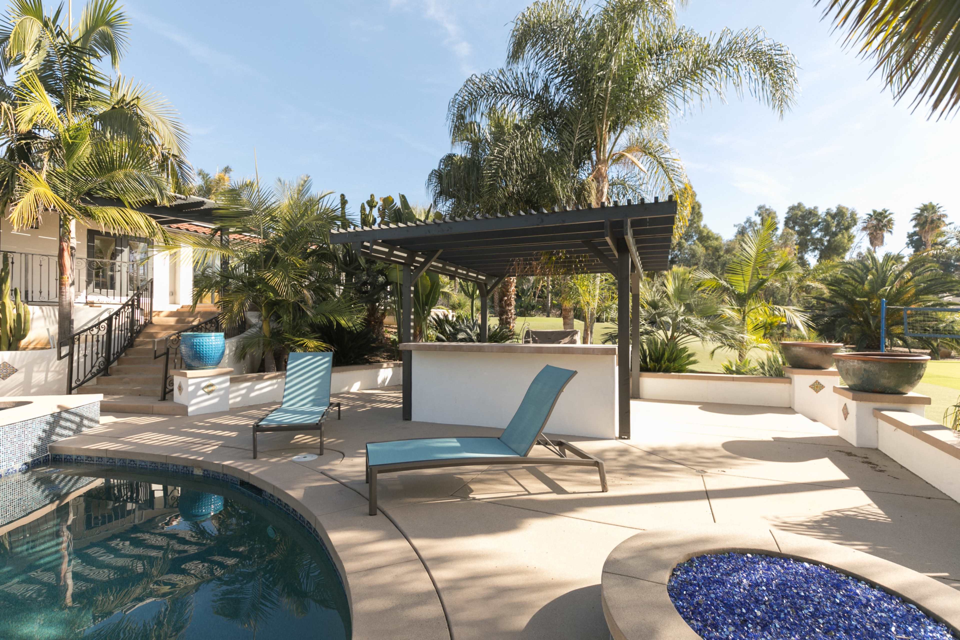The image features a landscaped outdoor area with a swimming pool, lounge chair, and a covered seating structure surrounded by palm trees and planters.