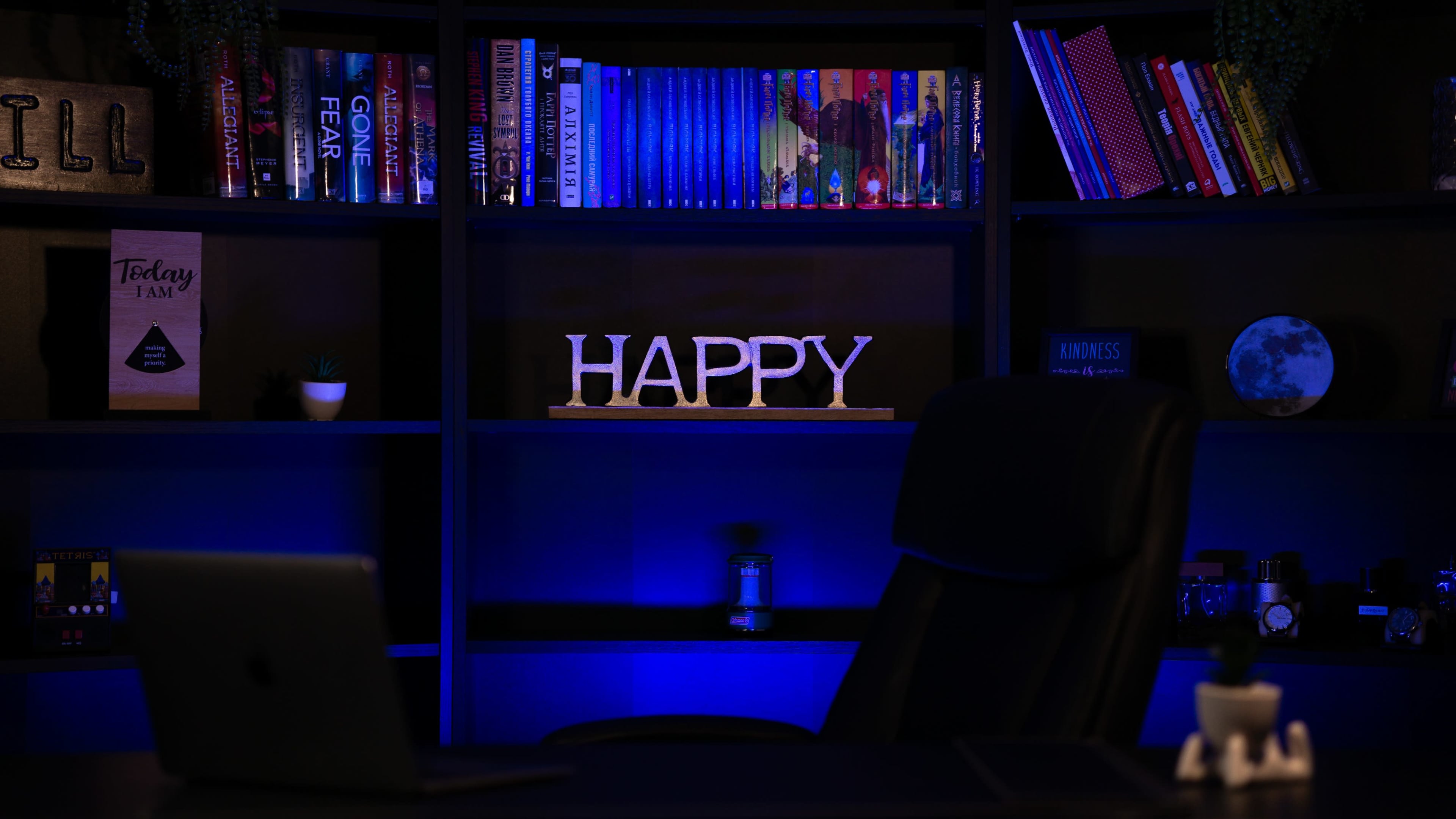 A black office chair is positioned in front of a shelf filled with colorful books, where a wooden sign spells "HAPPY" against a blue-lit background.