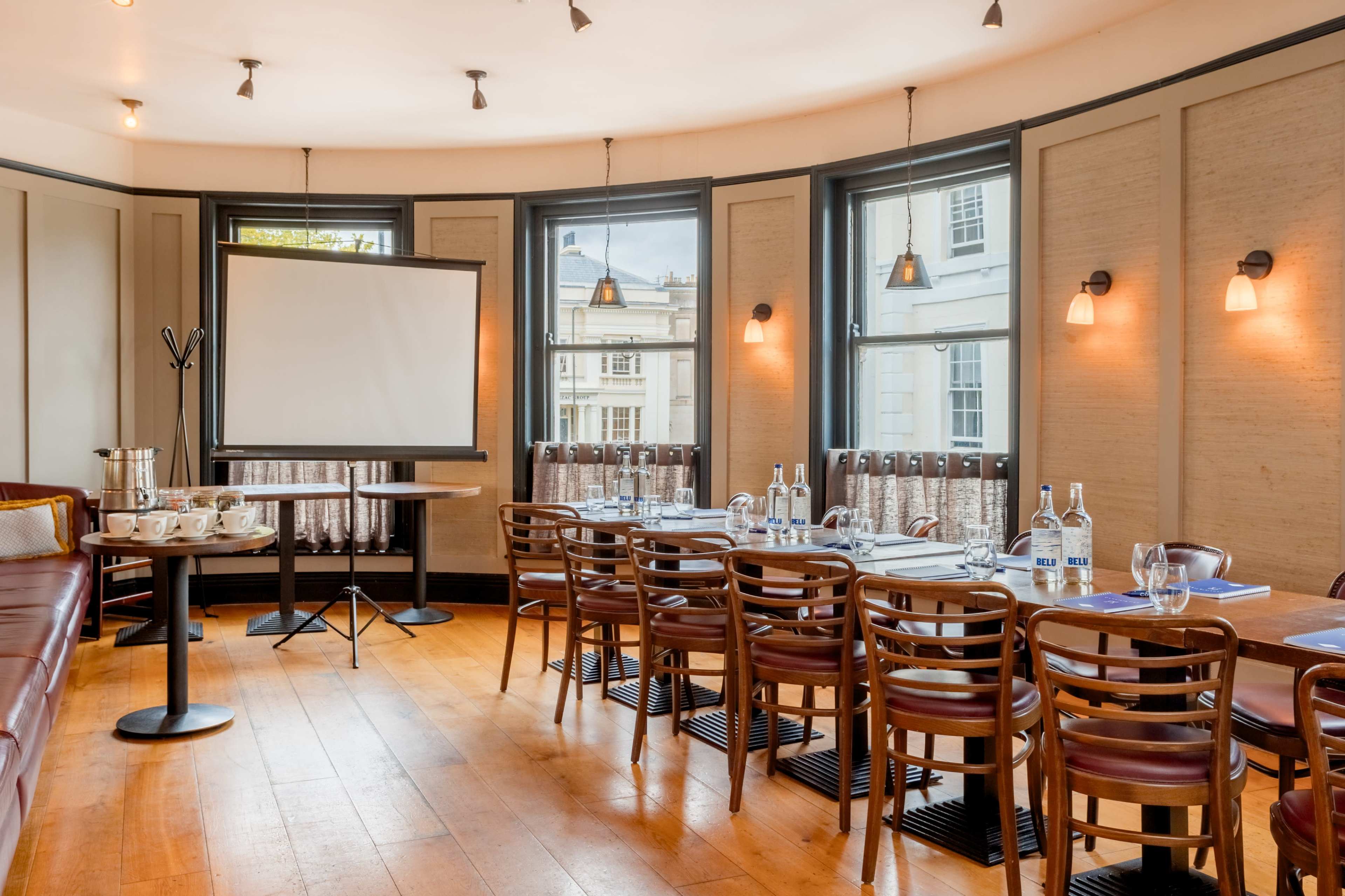 The image shows a conference room with a long table set up for a meeting, featuring chairs, a presentation screen, and large windows.