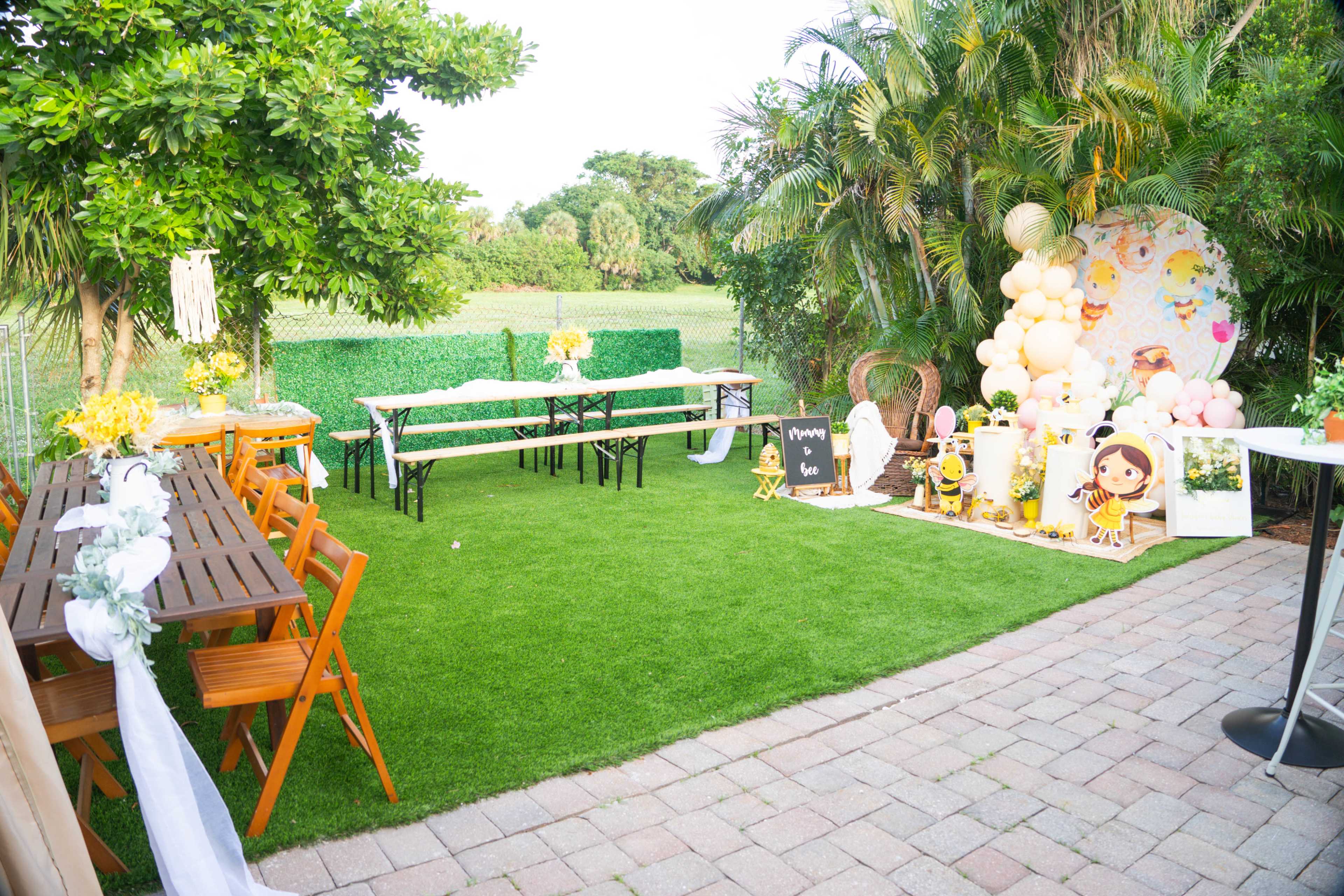 A backyard event space is set up with wooden tables and chairs, decorated with yellow flowers and a backdrop featuring balloons and a character display.