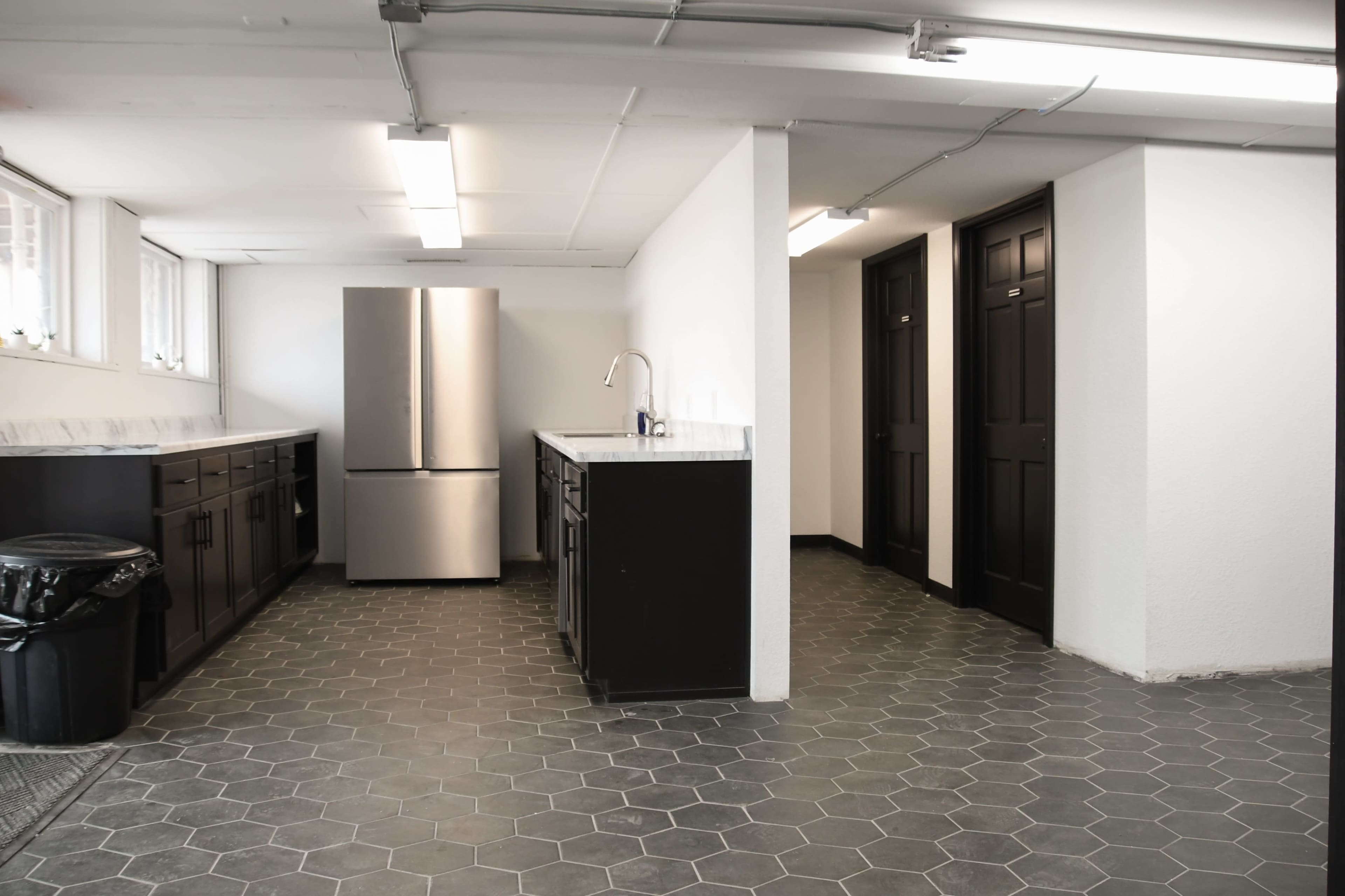 A spacious room features a stainless steel refrigerator, dark counters, and two closed doors along one wall, with a hexagonal tile floor.