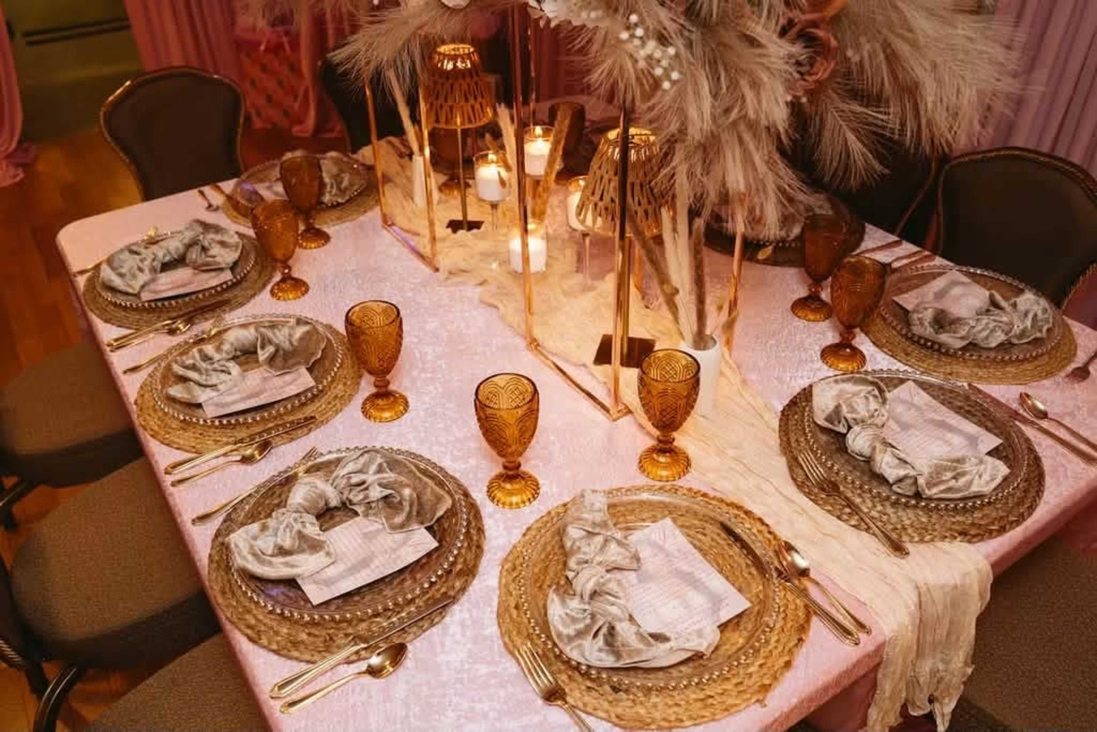 The image shows a elegantly set dining table surrounded by chairs, featuring woven placemats, decorative glassware, candles, and floral centerpieces.