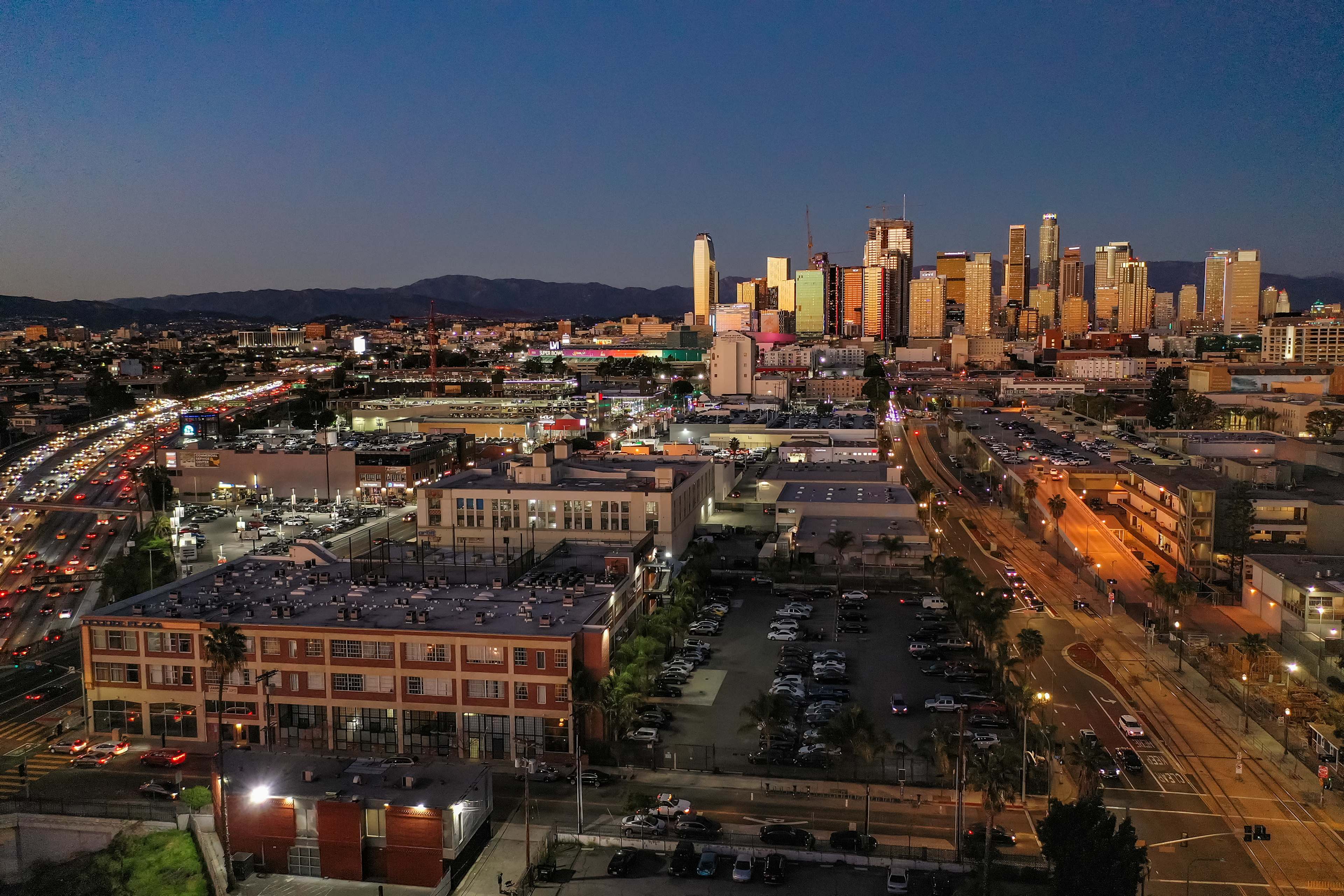 A cityscape of downtown Los Angeles at dusk, with skyscrapers illuminated against a darkening sky and busy roads filled with cars.