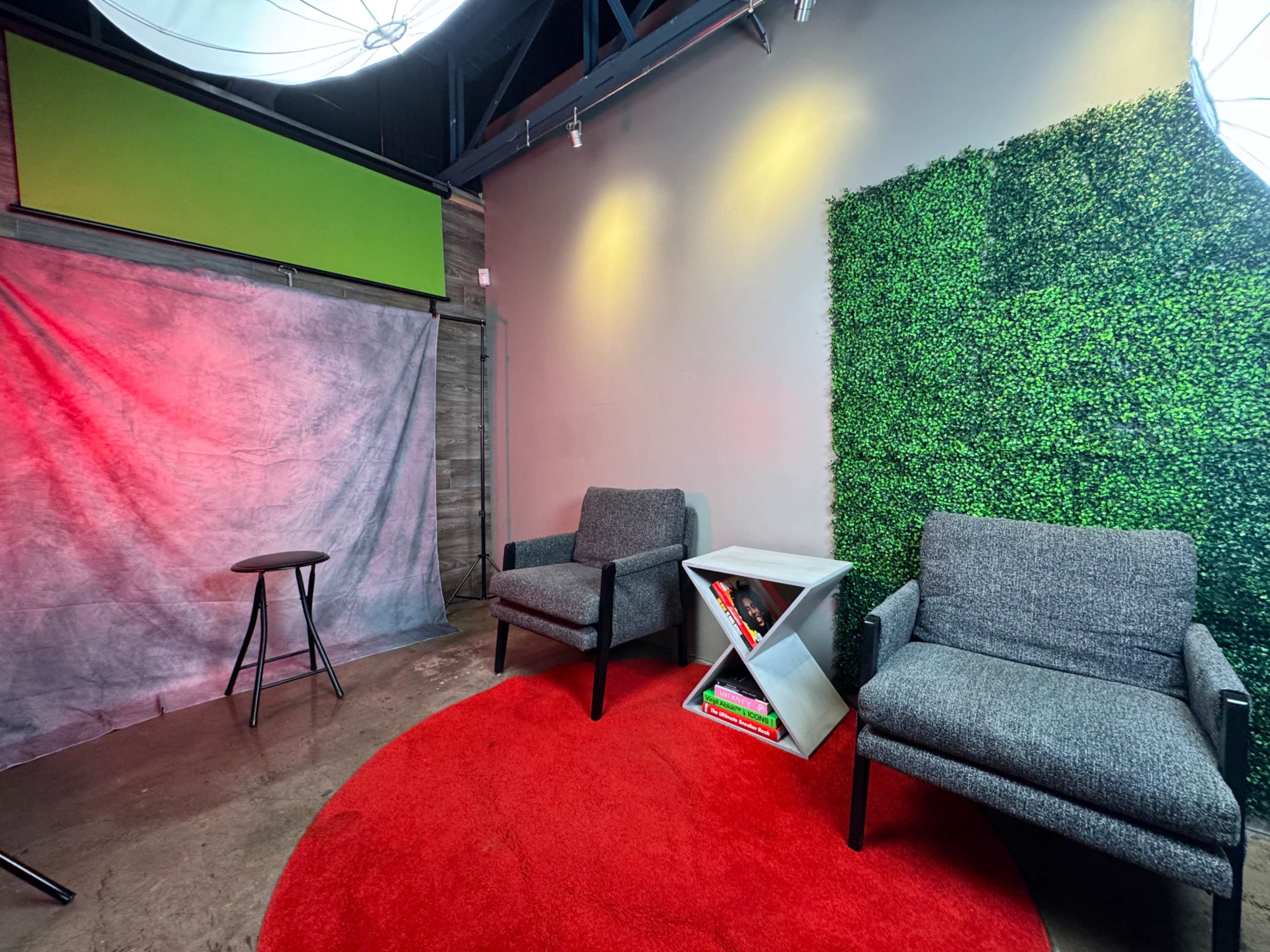 The image shows a studio space featuring two gray chairs, a round red carpet, a gray background, and a green wall covered in artificial foliage.
