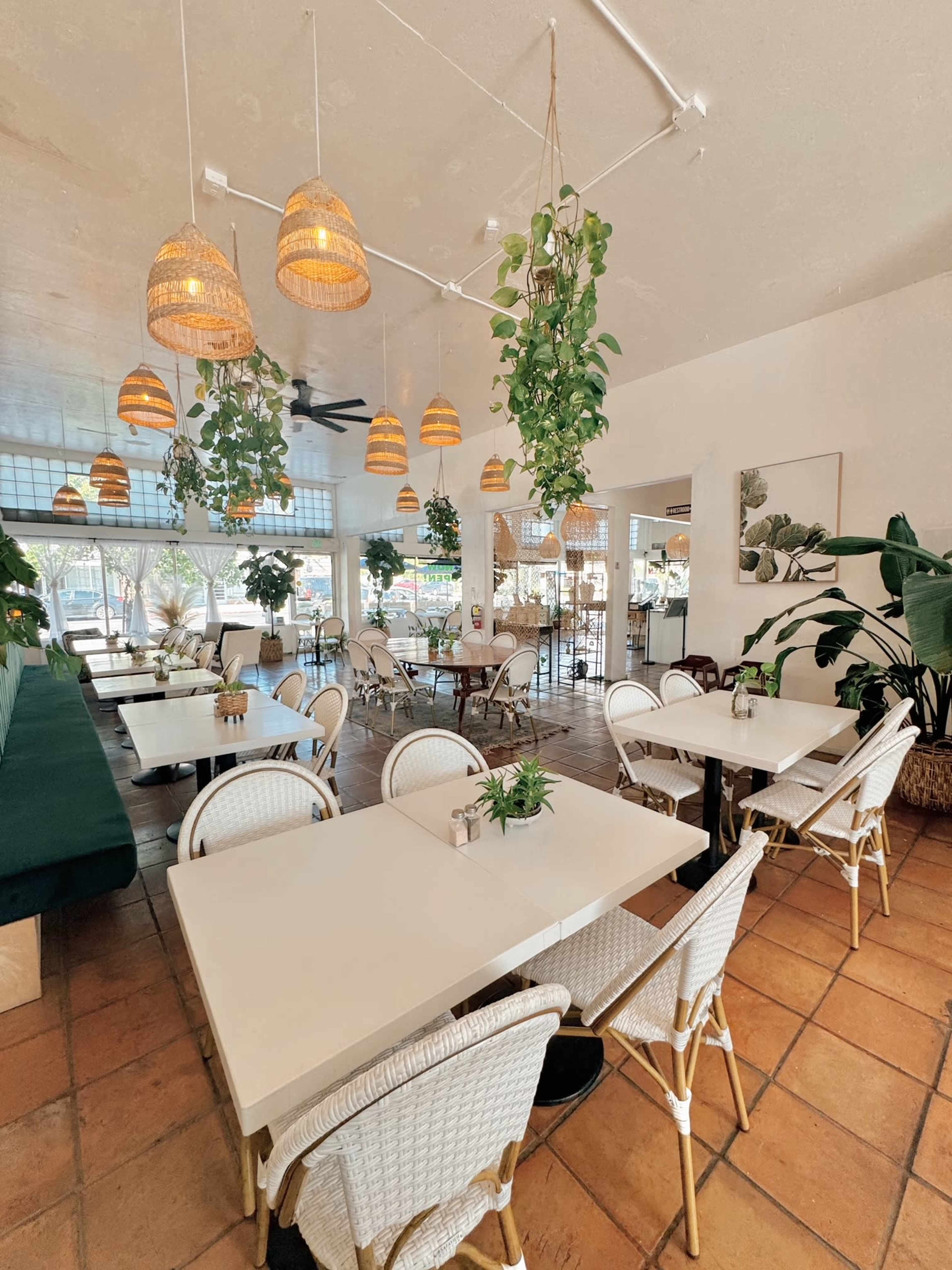 A bright, modern restaurant interior with white tables, woven chairs, hanging light fixtures, and potted plants.