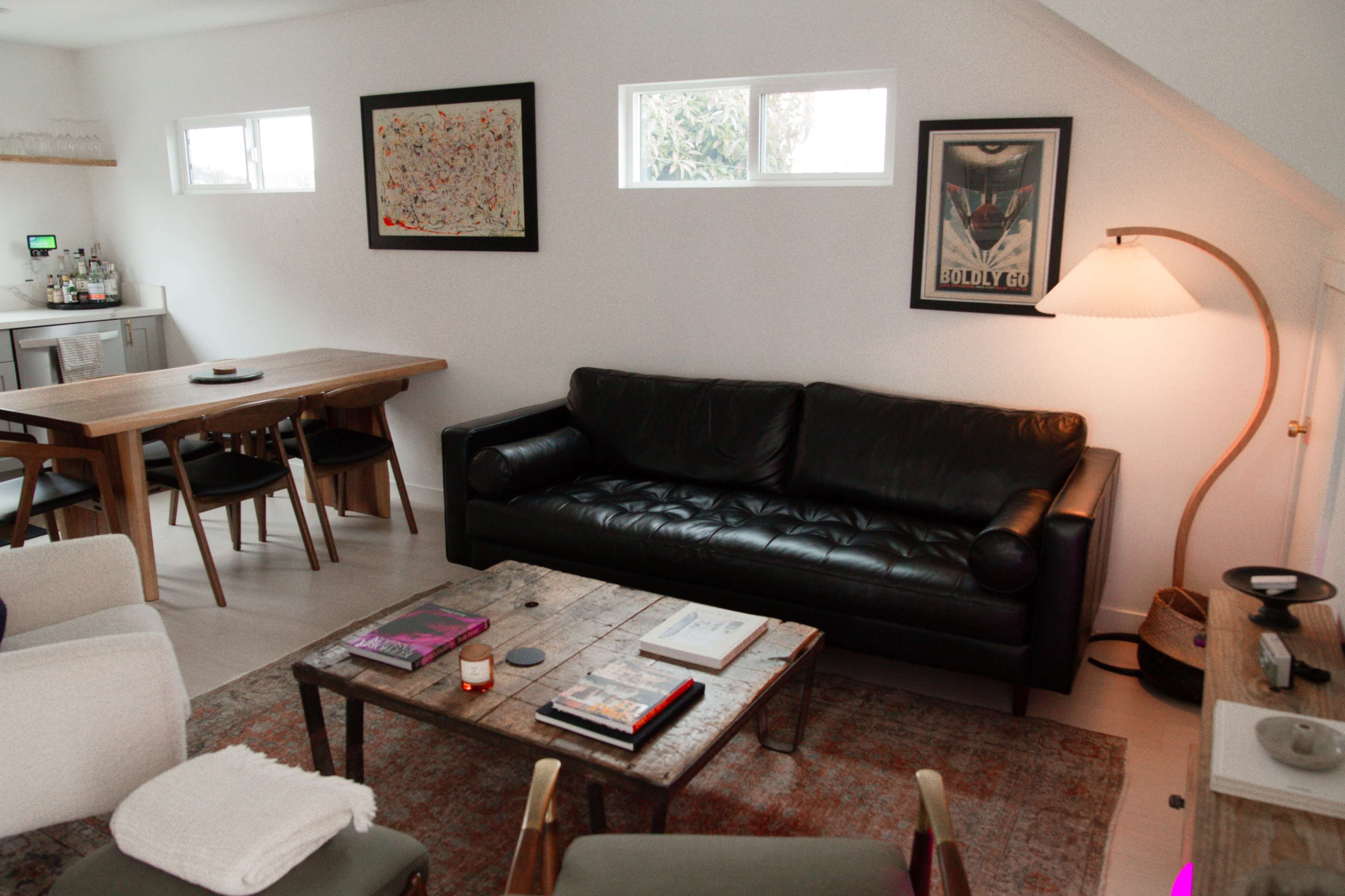 The image shows a modern living room with a black leather sofa, a wooden coffee table, a dining table, and artwork featured on the walls.