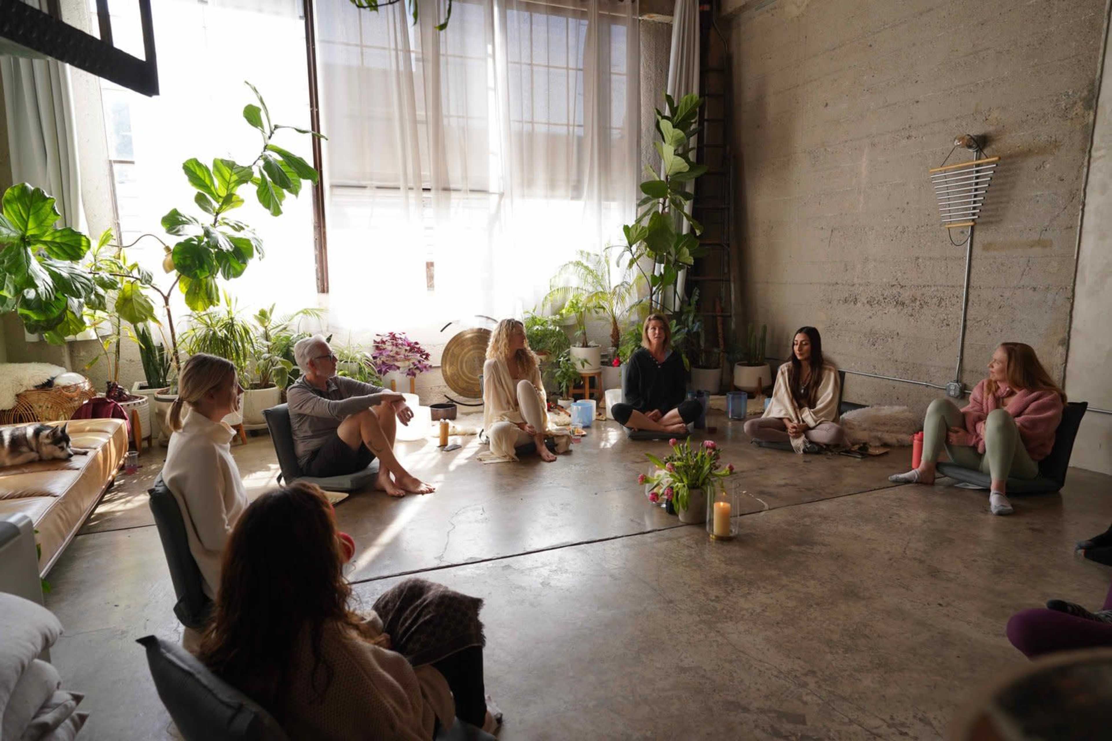 A group of people sit on the floor of a spacious, sunlit room surrounded by plants, candles, and scattered flowers.