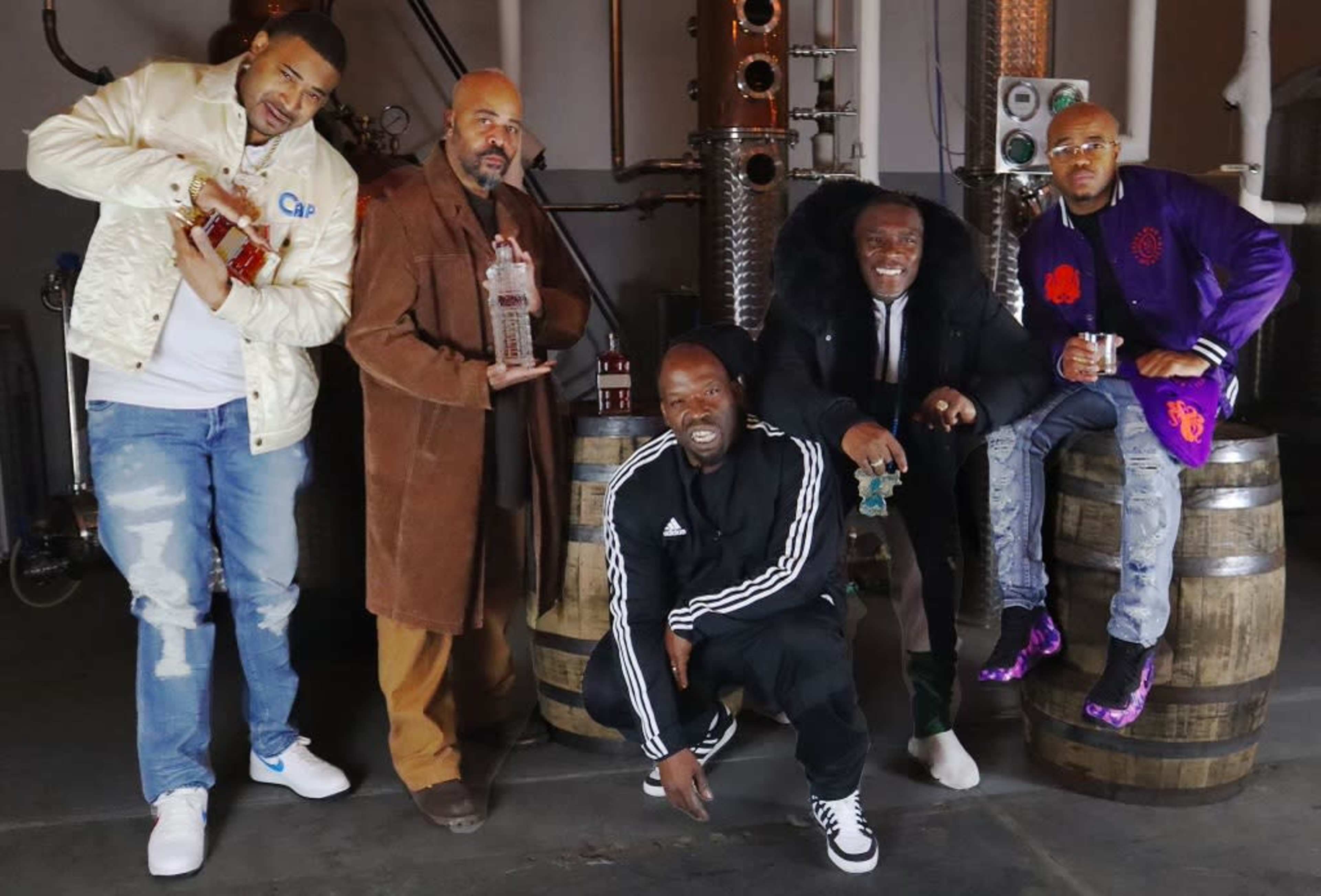Five men stand together in a distillery, holding bottles of liquor, with barrels in the background.