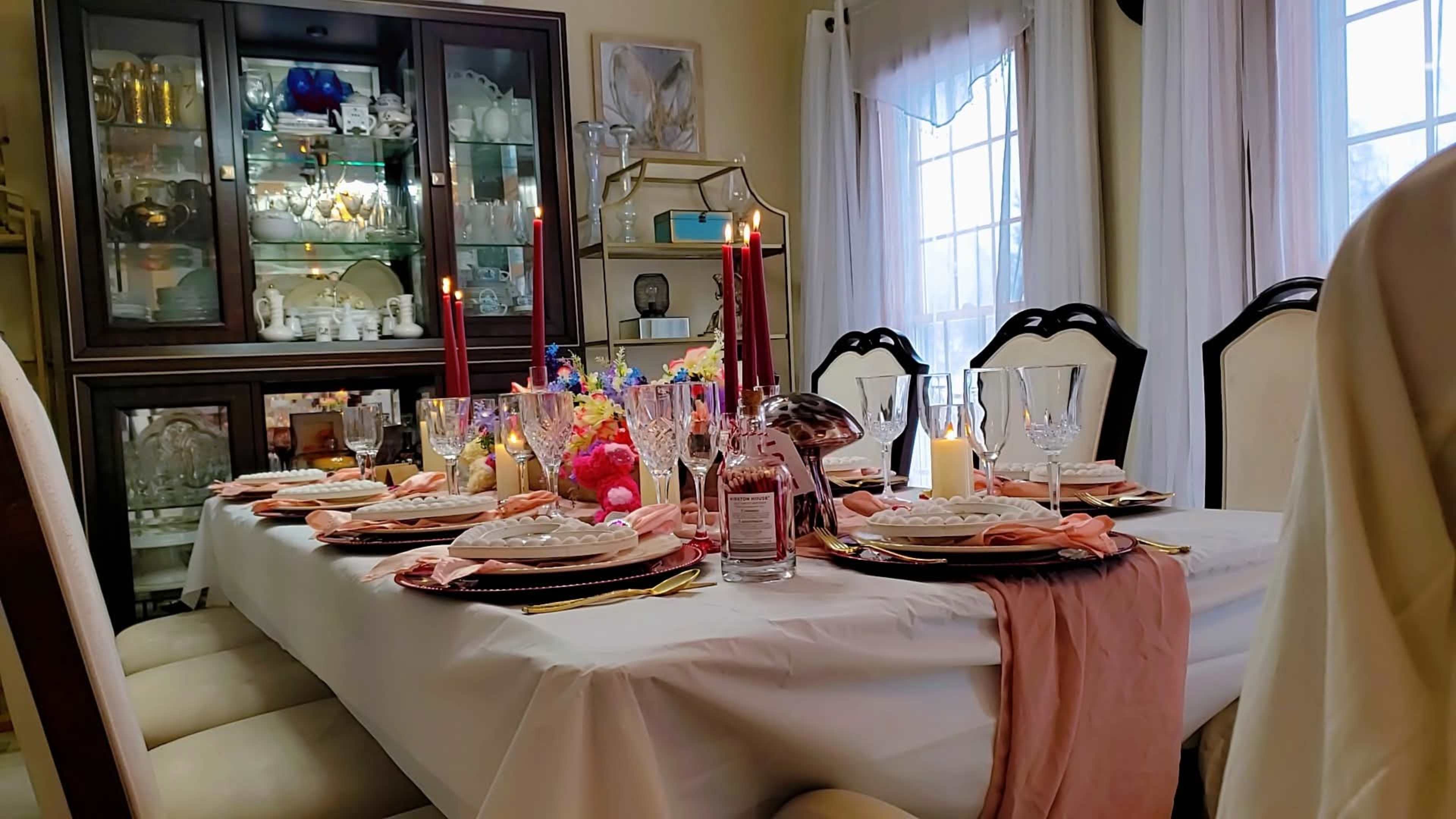 A dining table is set with elegant dishes, stemware, and candles, surrounded by light-colored chairs and a display cabinet in the background.