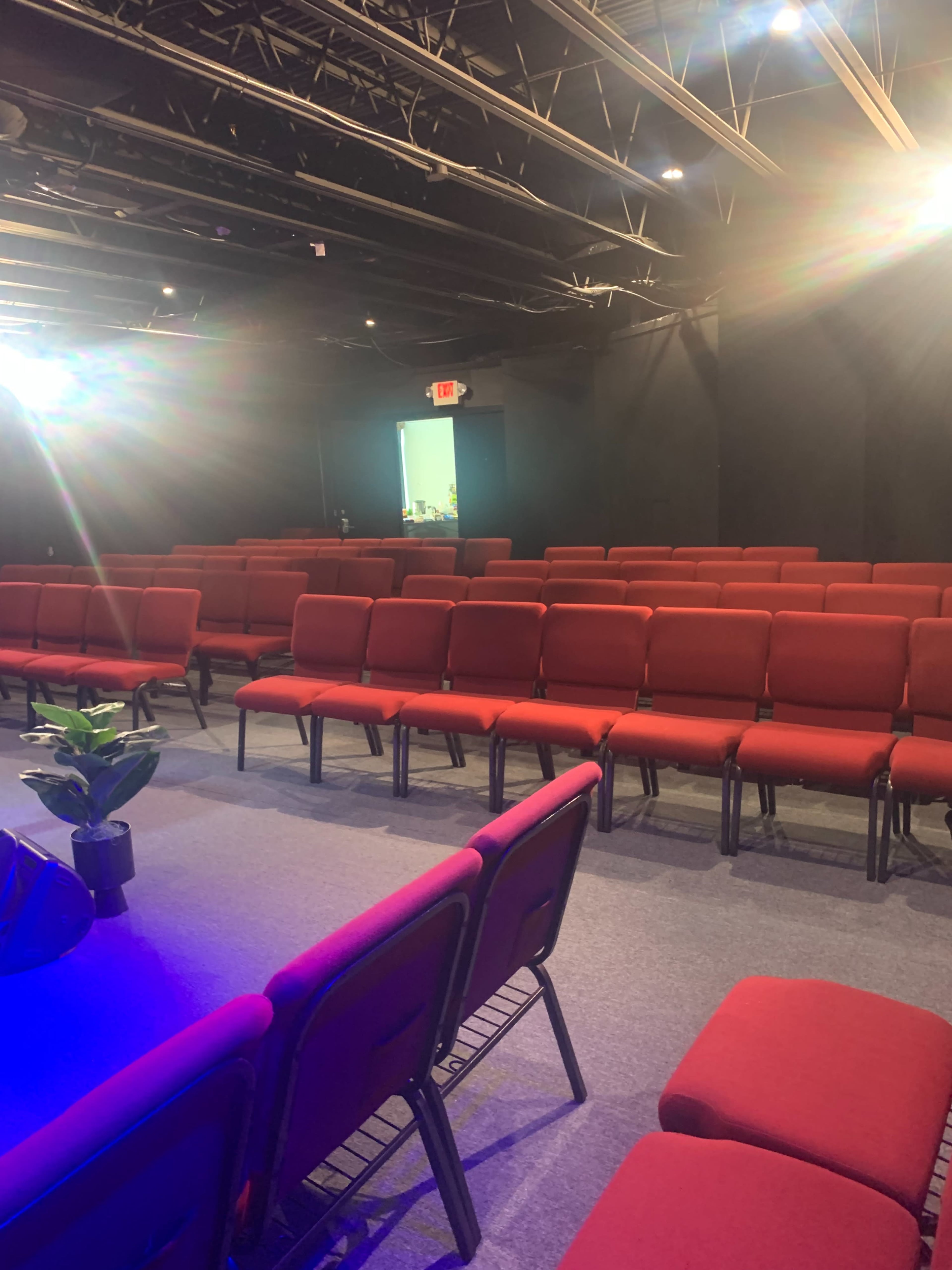 The image shows a row of red chairs arranged in a dimly lit auditorium space, with bright lights illuminating the area.