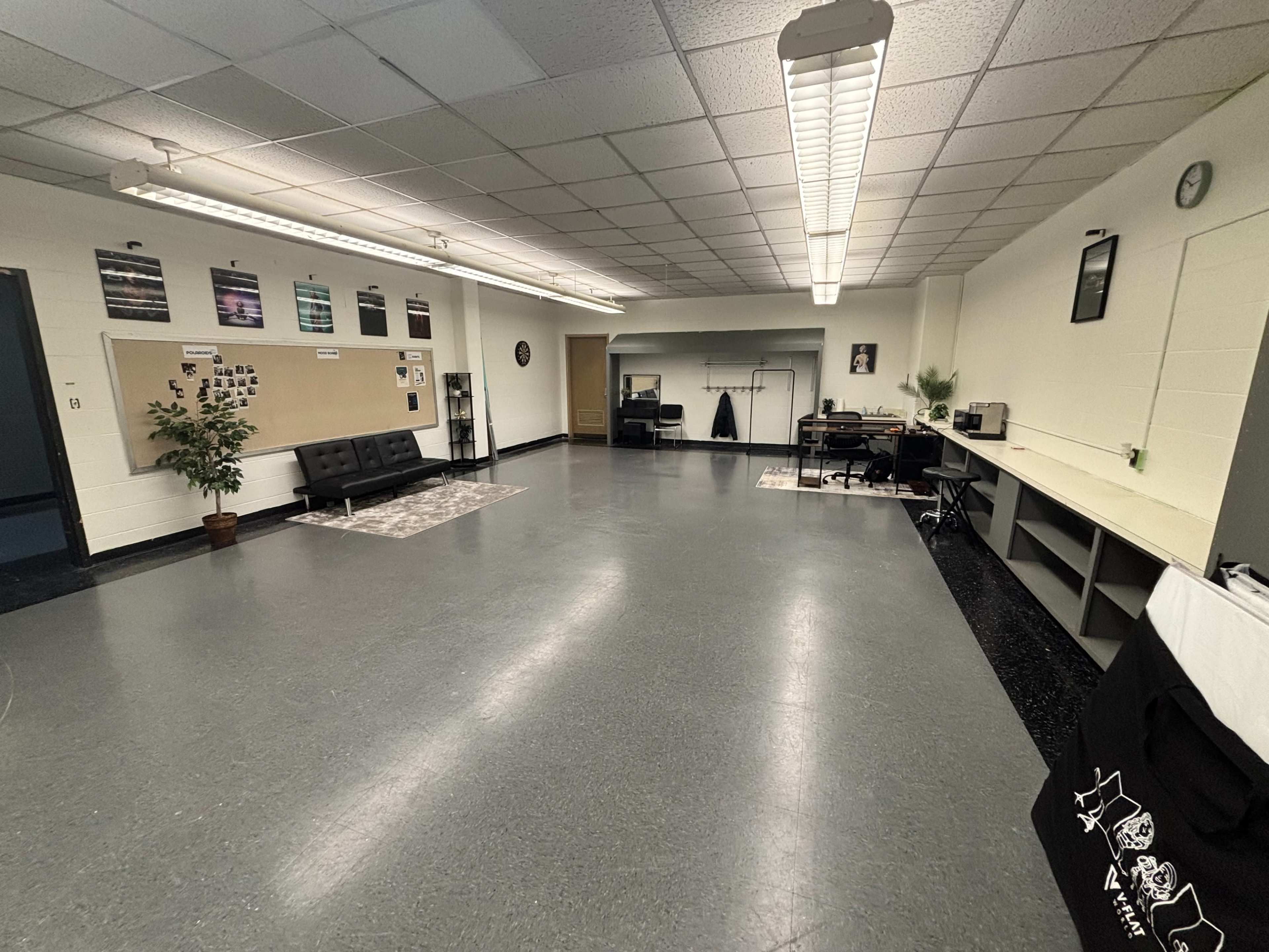 The image shows a spacious, well-lit room featuring a black futon, a plant in a pot, a large table, and various items along the walls, including posters and a clock.