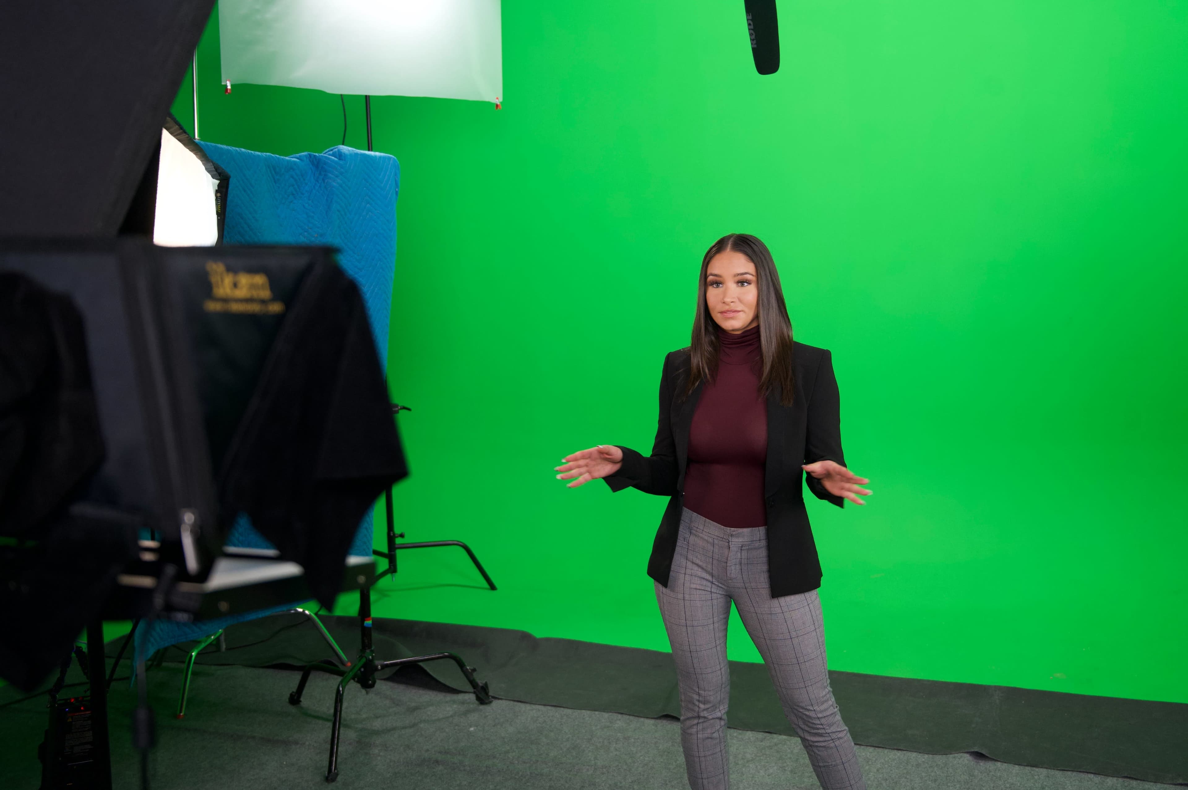 A woman stands in front of a green screen, gesturing with her hands while preparing for a presentation, with studio lighting and equipment visible.