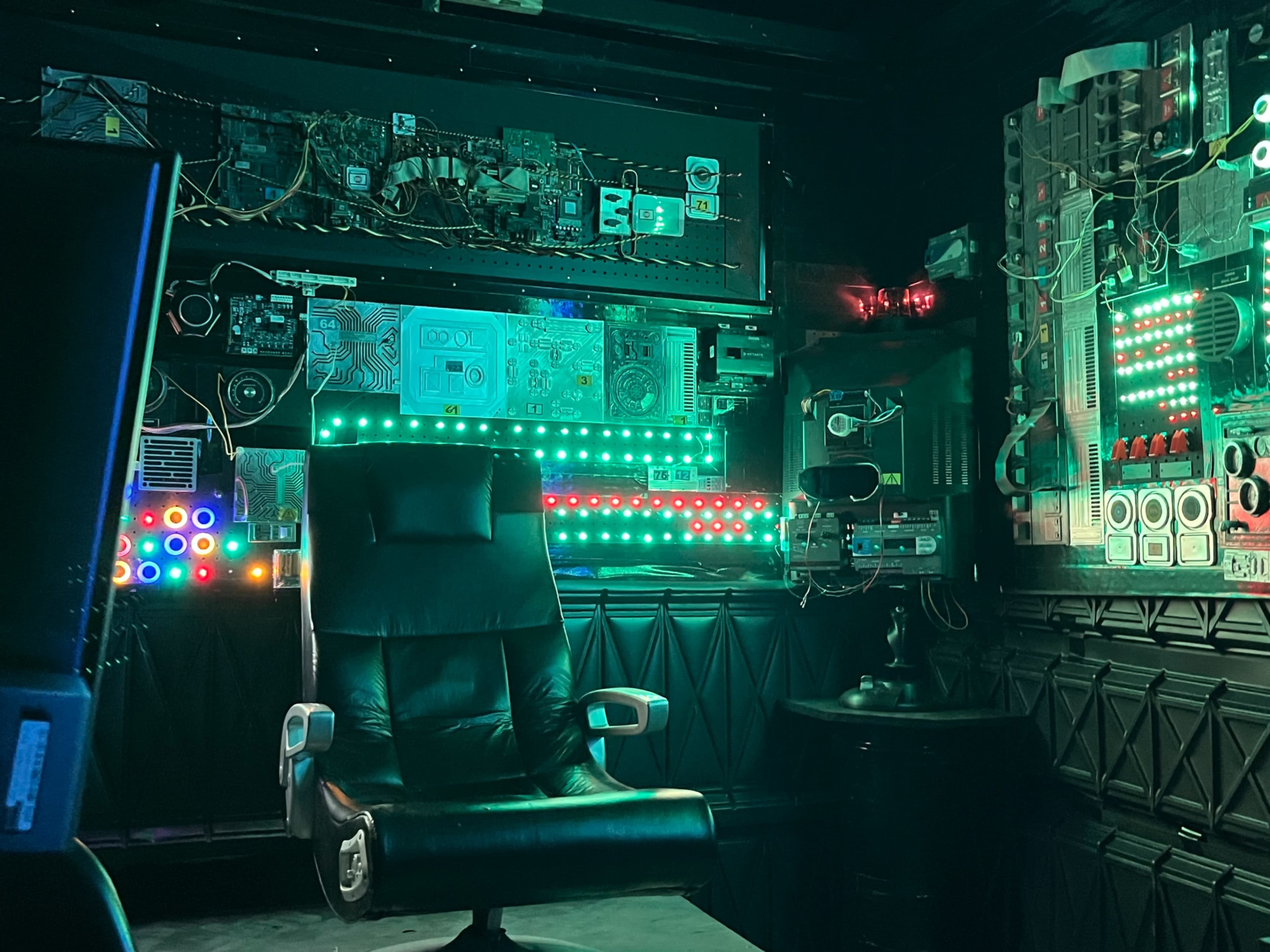The image shows a dark, high-tech room filled with electronic equipment and illuminated with colorful LED lights, featuring a central black leather chair.
