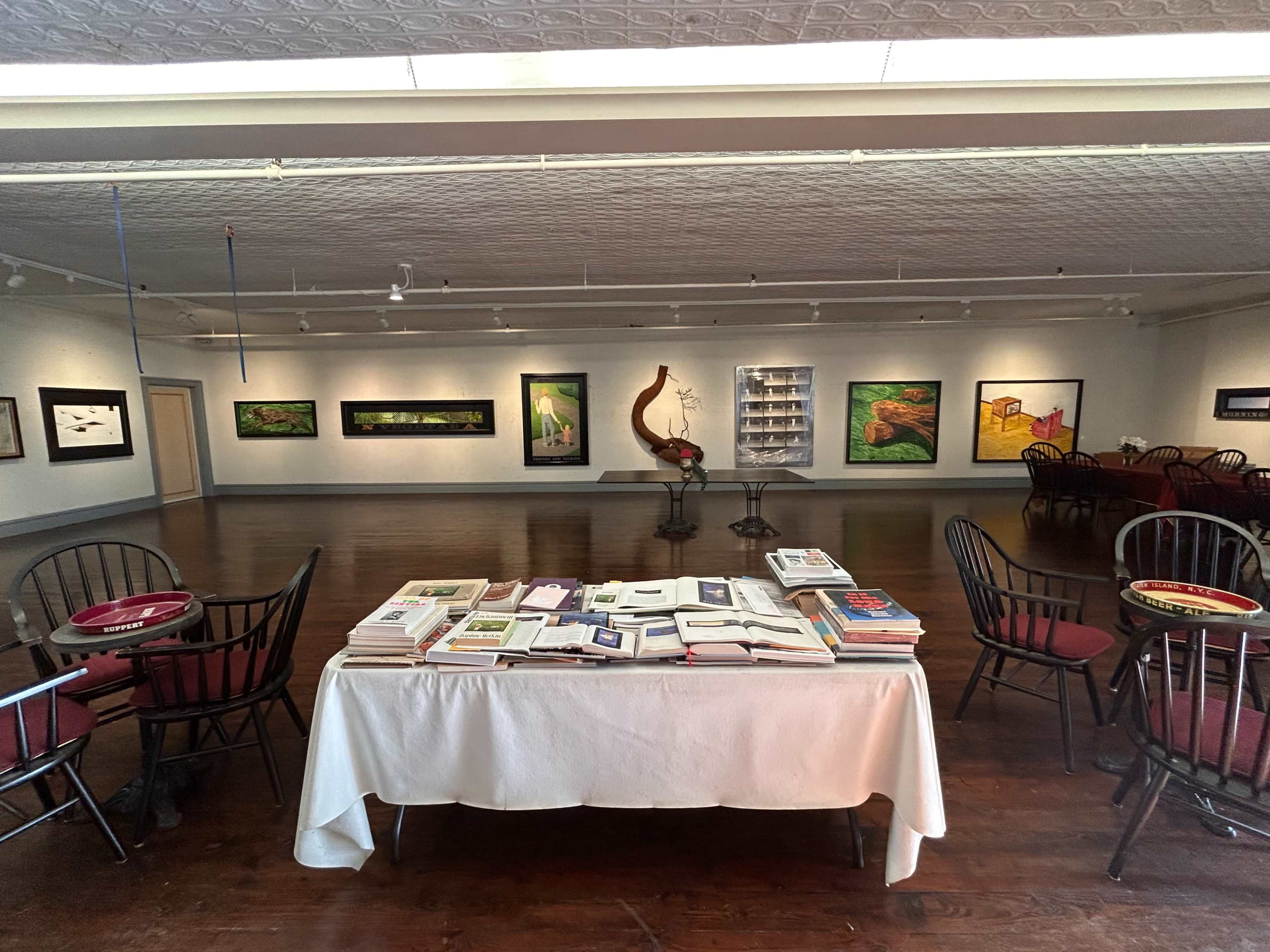 The image shows an art gallery featuring a table covered with books and a number of framed artworks displayed on the walls.