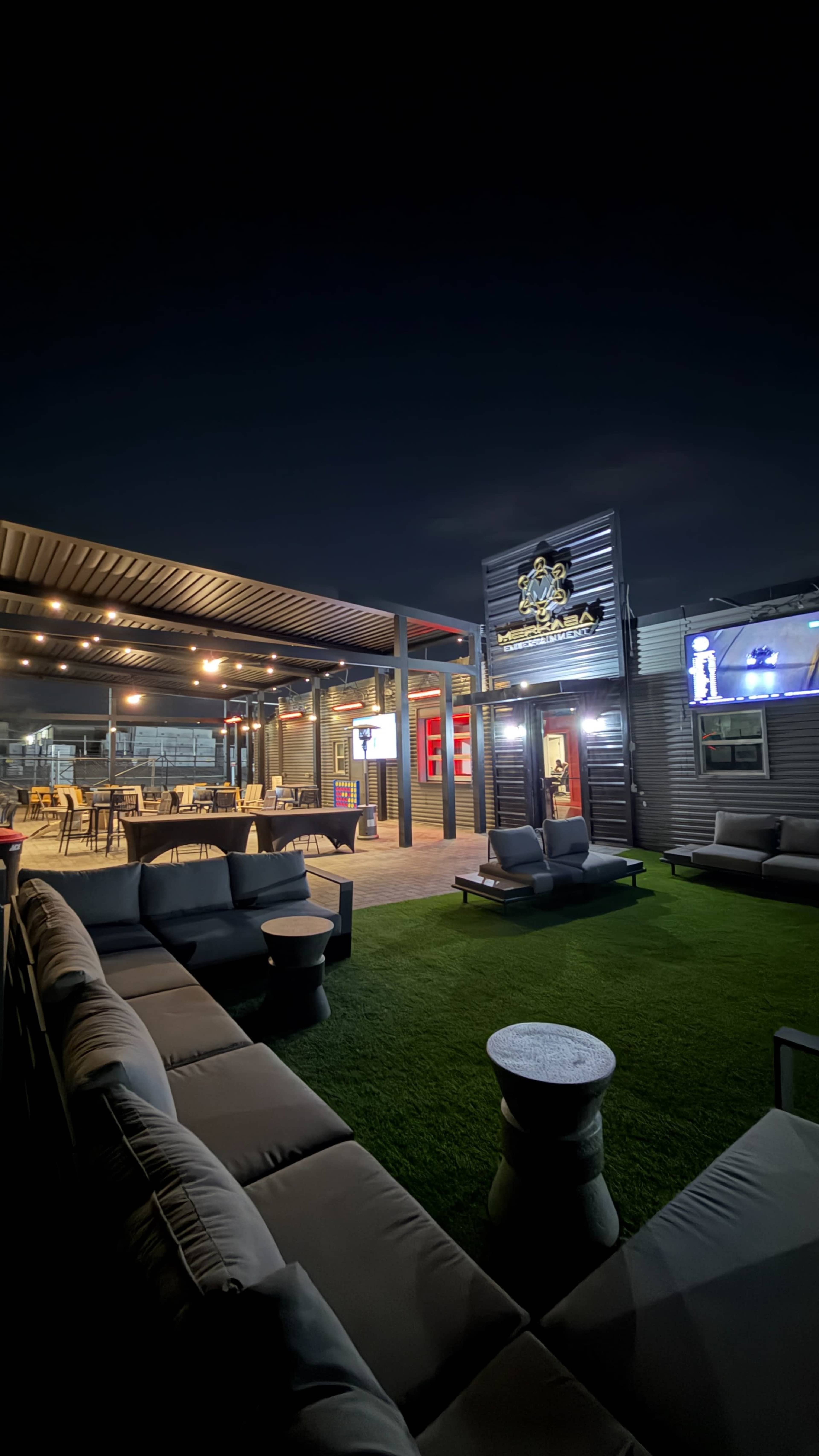 The image shows a modern rooftop lounge area with outdoor seating, ambient lighting, and a bar in the background.