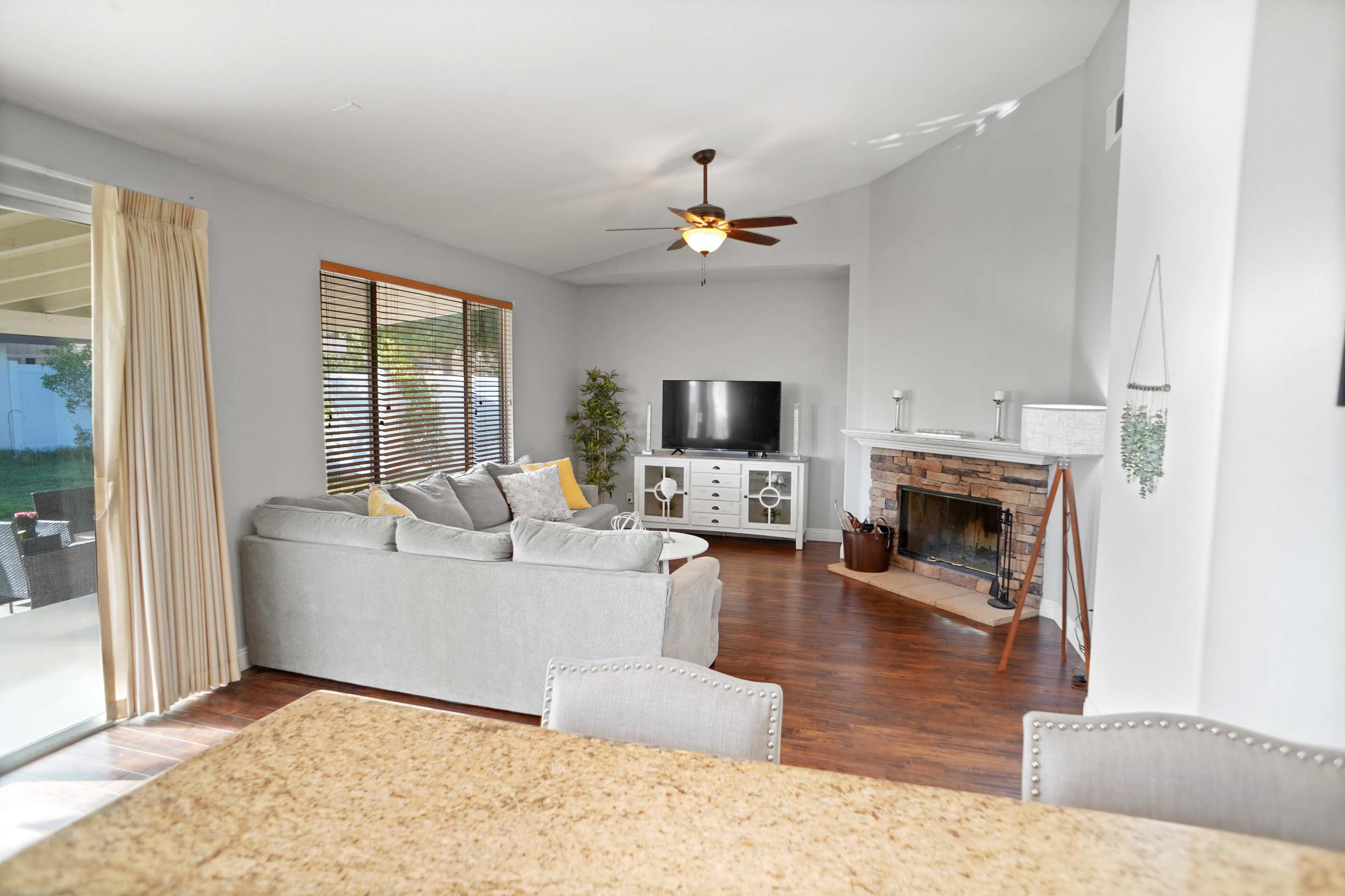 The image shows a living room with a gray sofa, a television on a white entertainment center, and a brick fireplace, all illuminated by natural light from a nearby window.