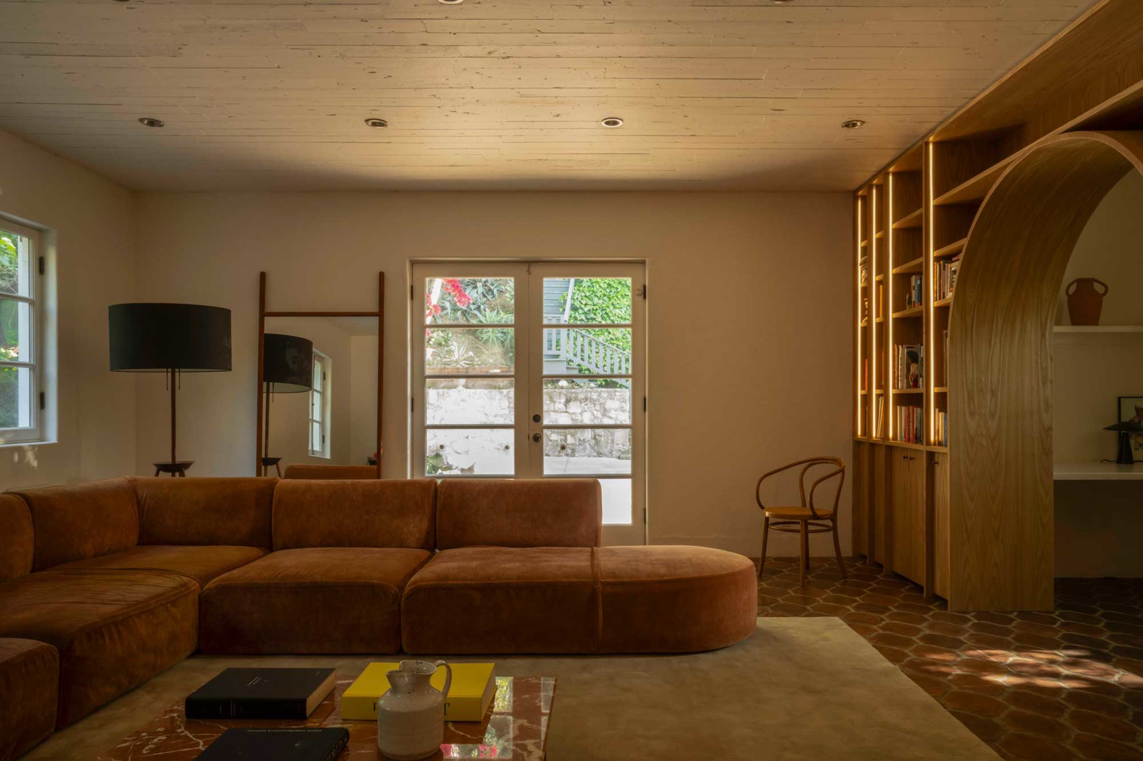 A spacious living room features a curved brown sofa, wooden bookshelves, and large glass doors opening to a garden.