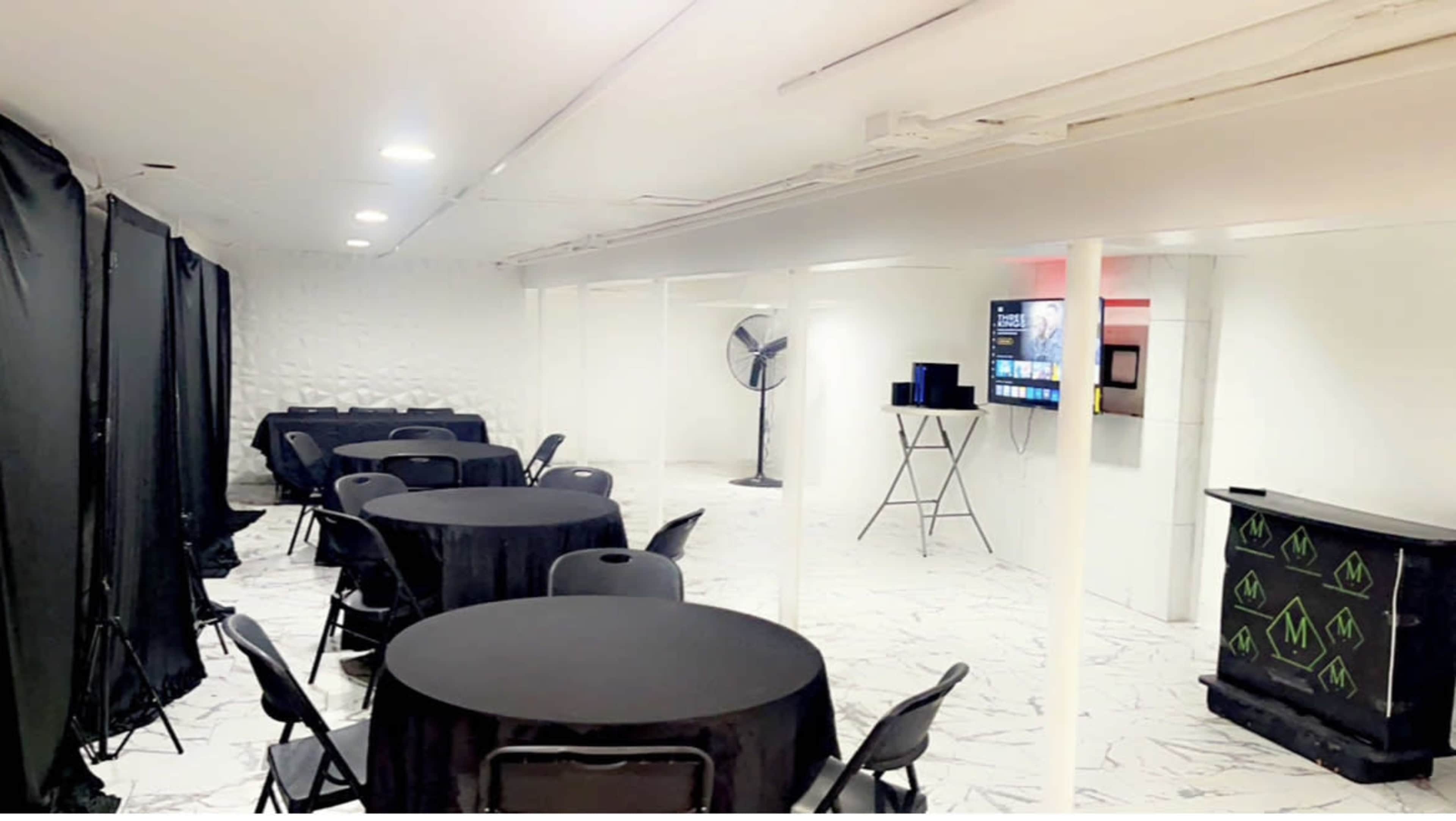 A brightly lit, minimalist event space with black tables and chairs arranged throughout, featuring a television mounted on the wall and a fan in the corner.