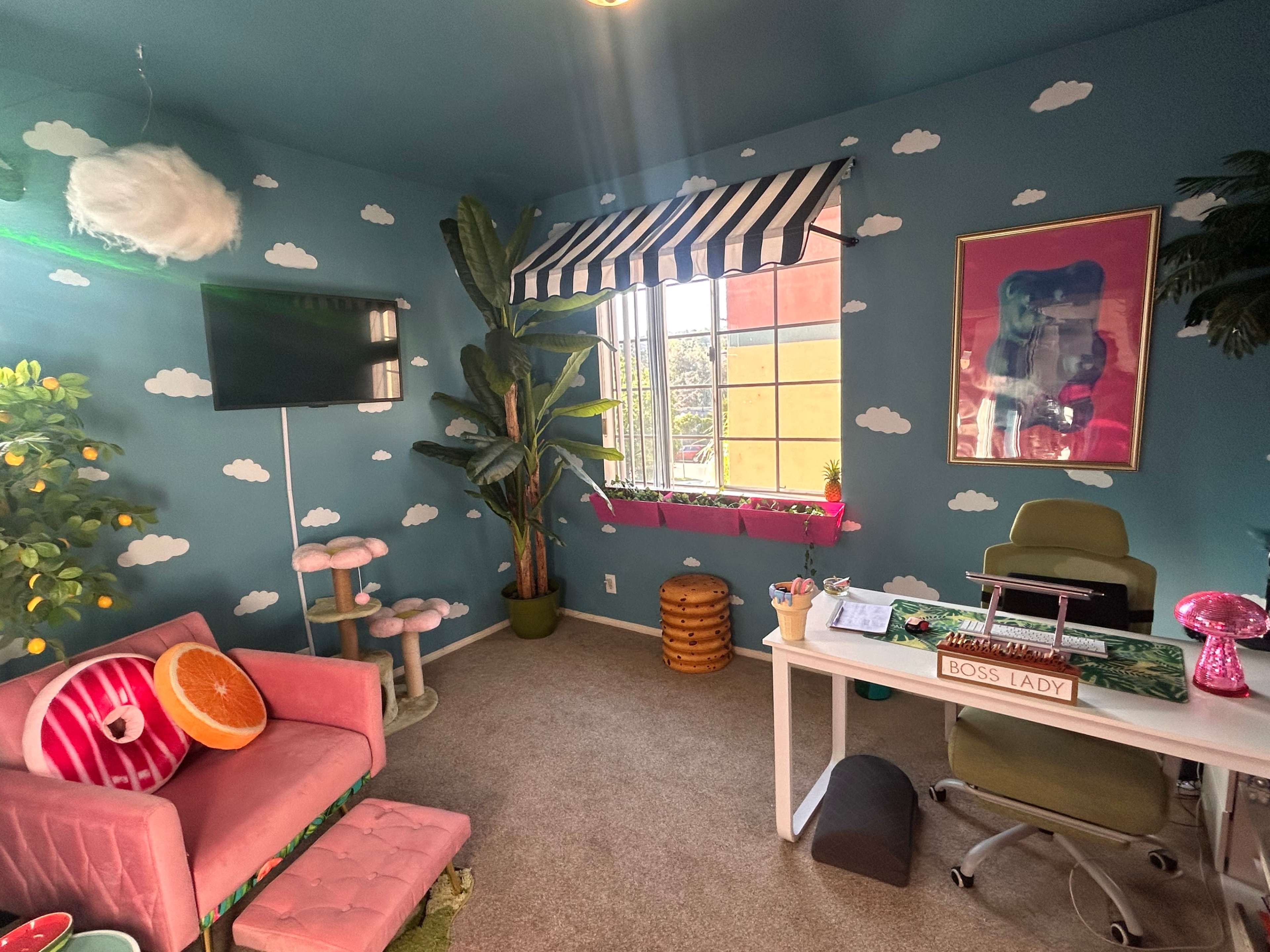The image shows a brightly colored home office with a teal wall decorated with cloud motifs, a desk with a computer, and plush furniture including a pink couch and cat tree.