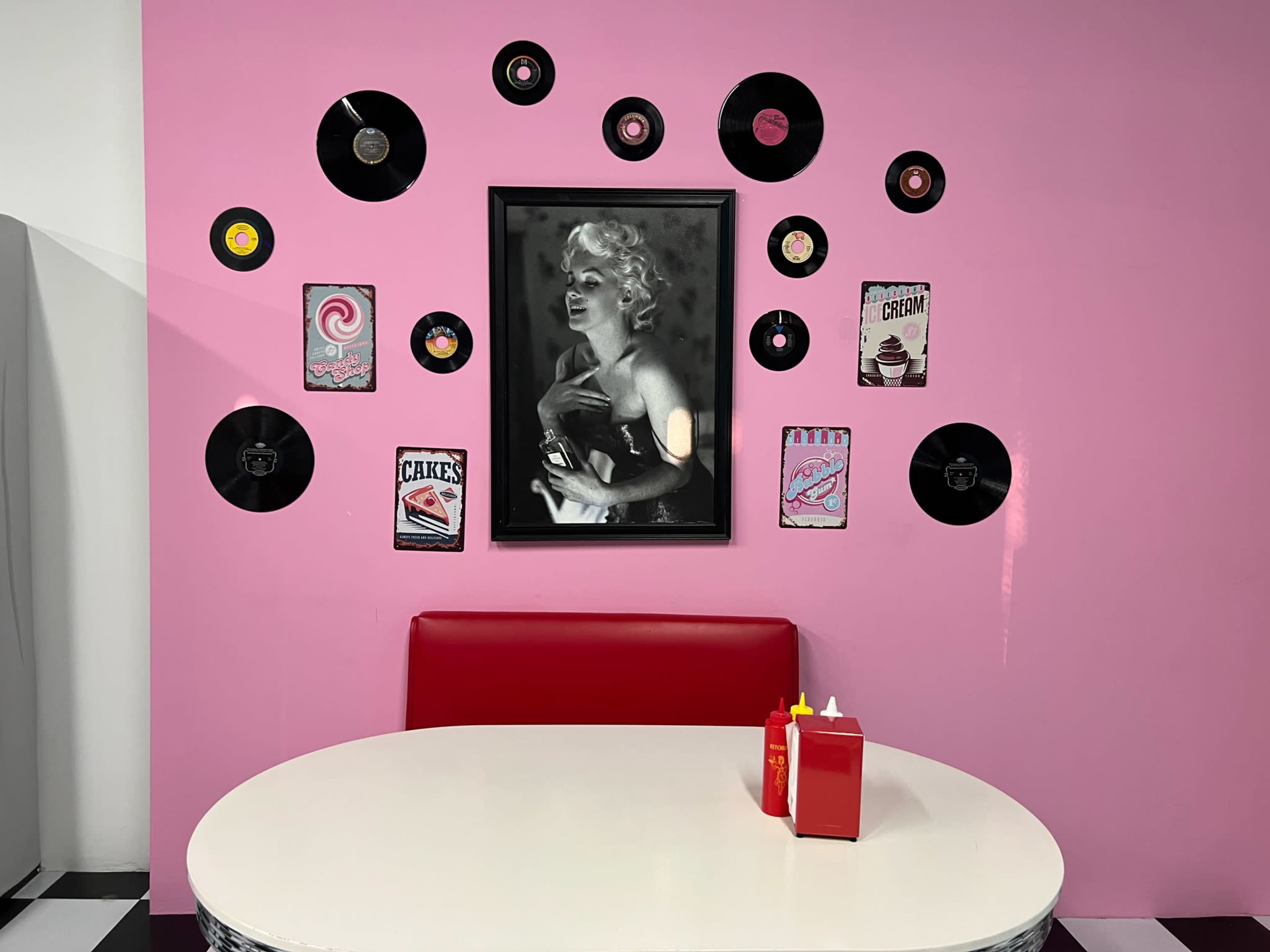 The scene features a round table with a red booth in front of a pink wall decorated with vintage records and retro ice cream-themed posters, including a prominent black-and-white photo of a woman.