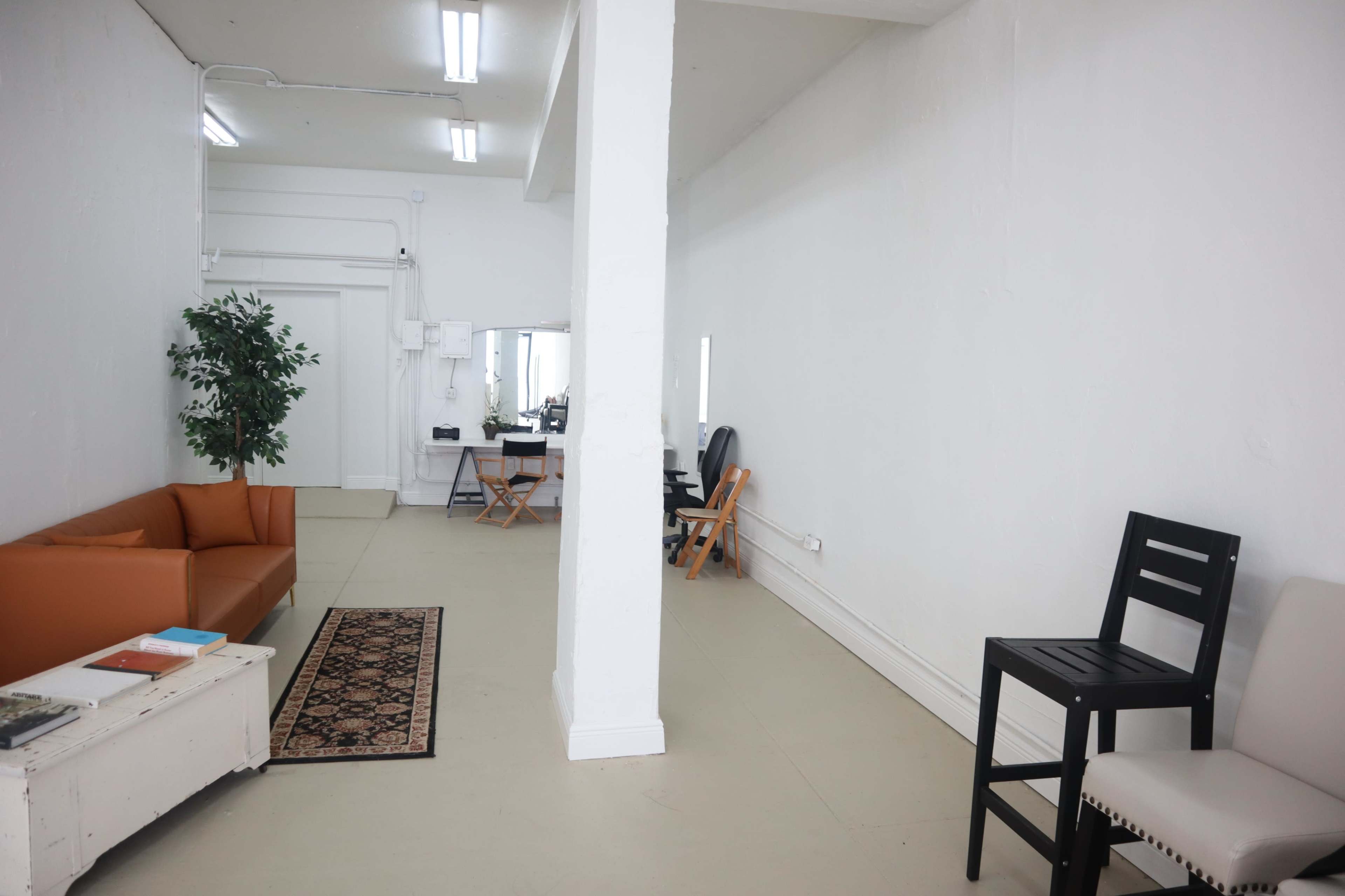 The image shows a spacious, minimalistic room with white walls, a brown sofa, a small rug, and a few chairs arranged on either side of an interior dividing column.