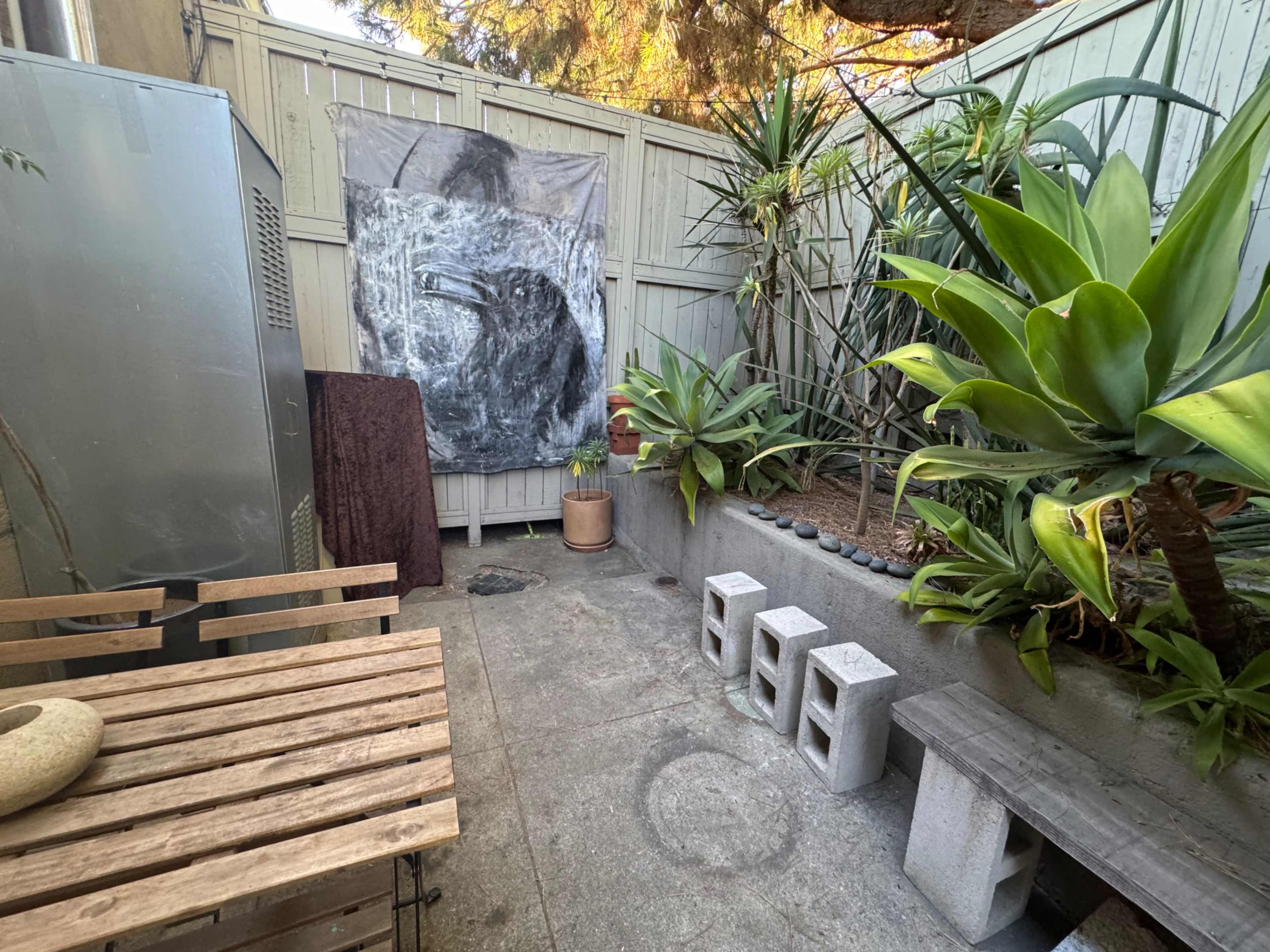 A small outdoor space with a concrete floor, a wooden table and benches, plants along the walls, and a large crow artwork hanging on the fence.