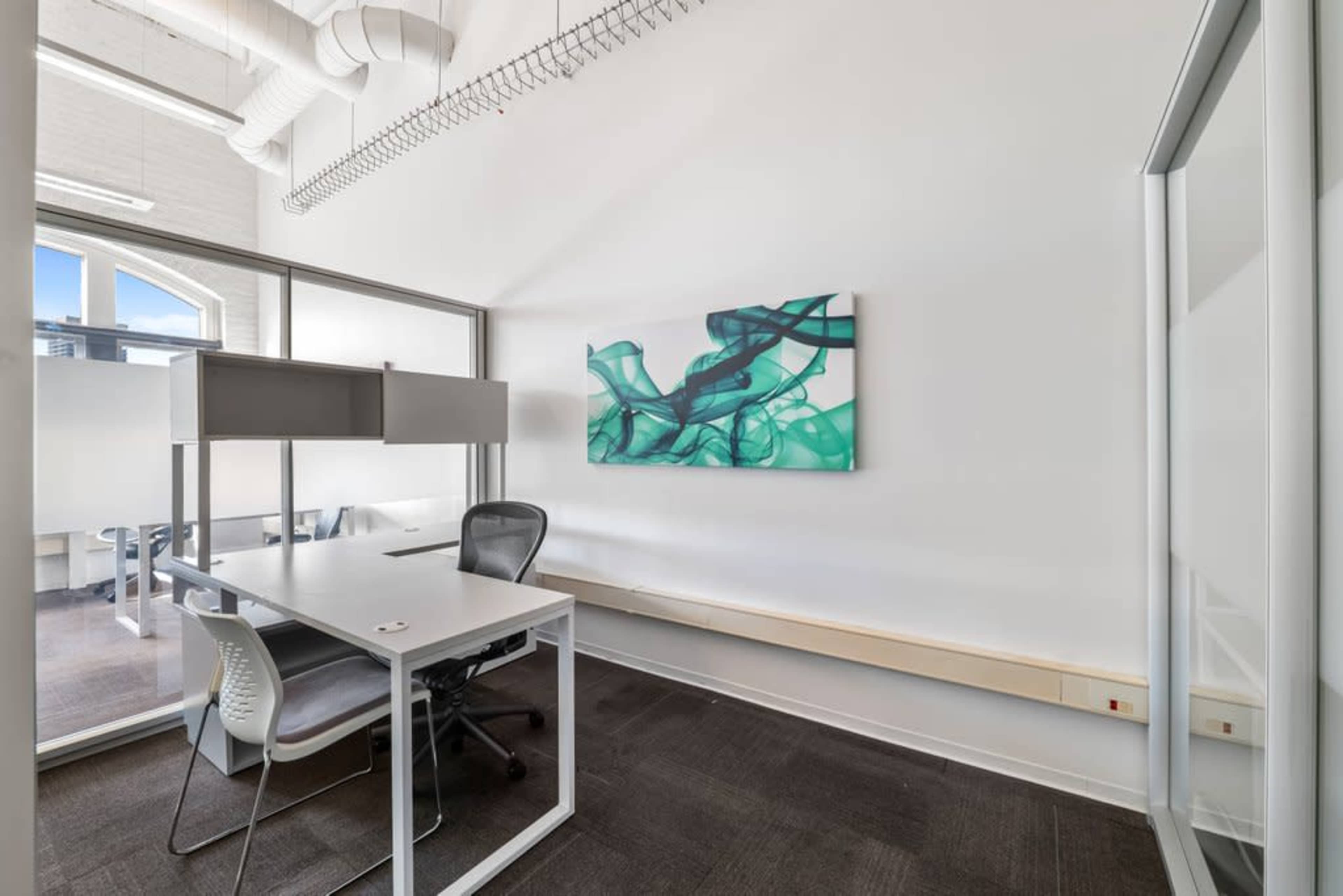 The image shows a modern office space featuring a white table with two chairs, a piece of wall art, and large windows with natural light.