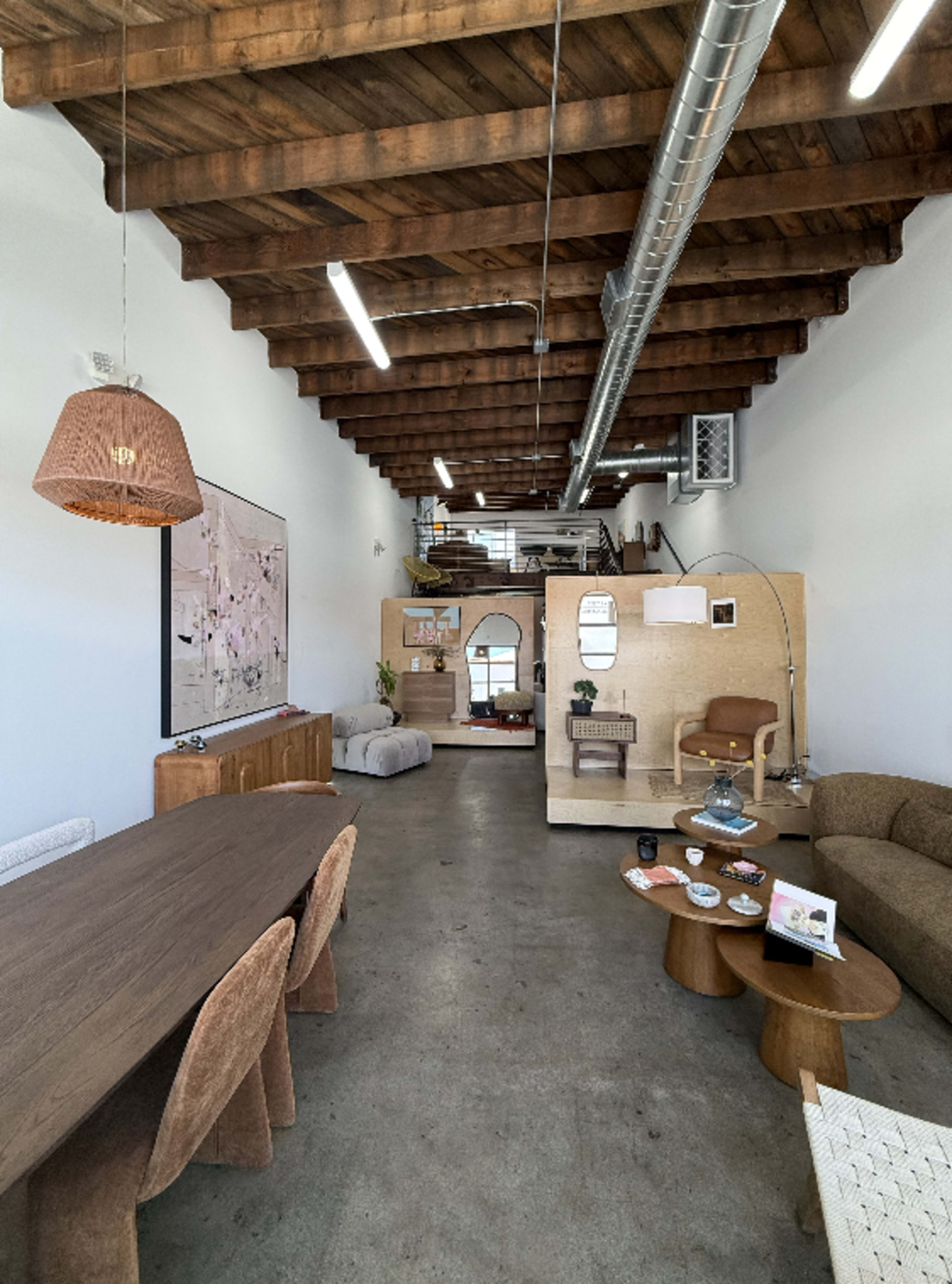 The image shows a spacious interior with a long wooden dining table, modern furniture, and a high ceiling featuring exposed beams.