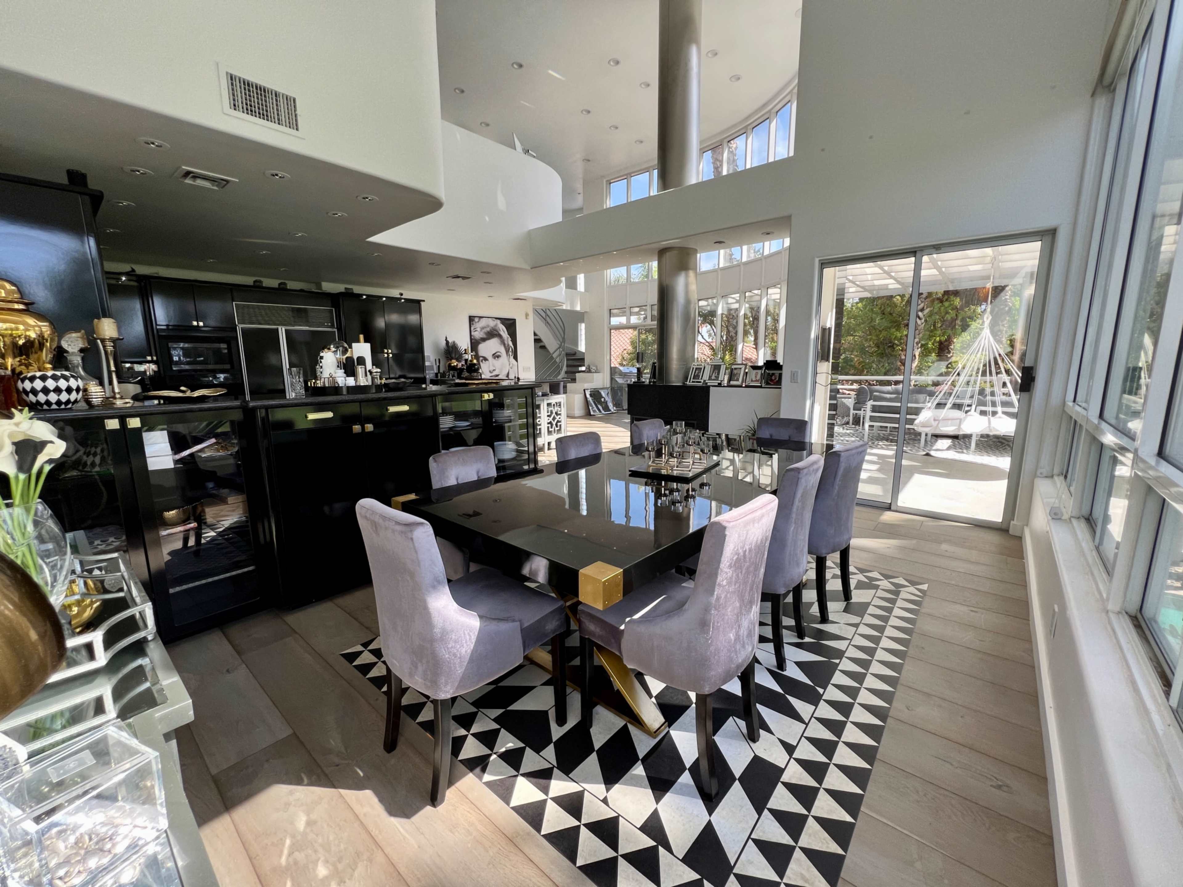 A modern dining area features a large black table surrounded by purple chairs, with an open kitchen and large windows that provide natural light.