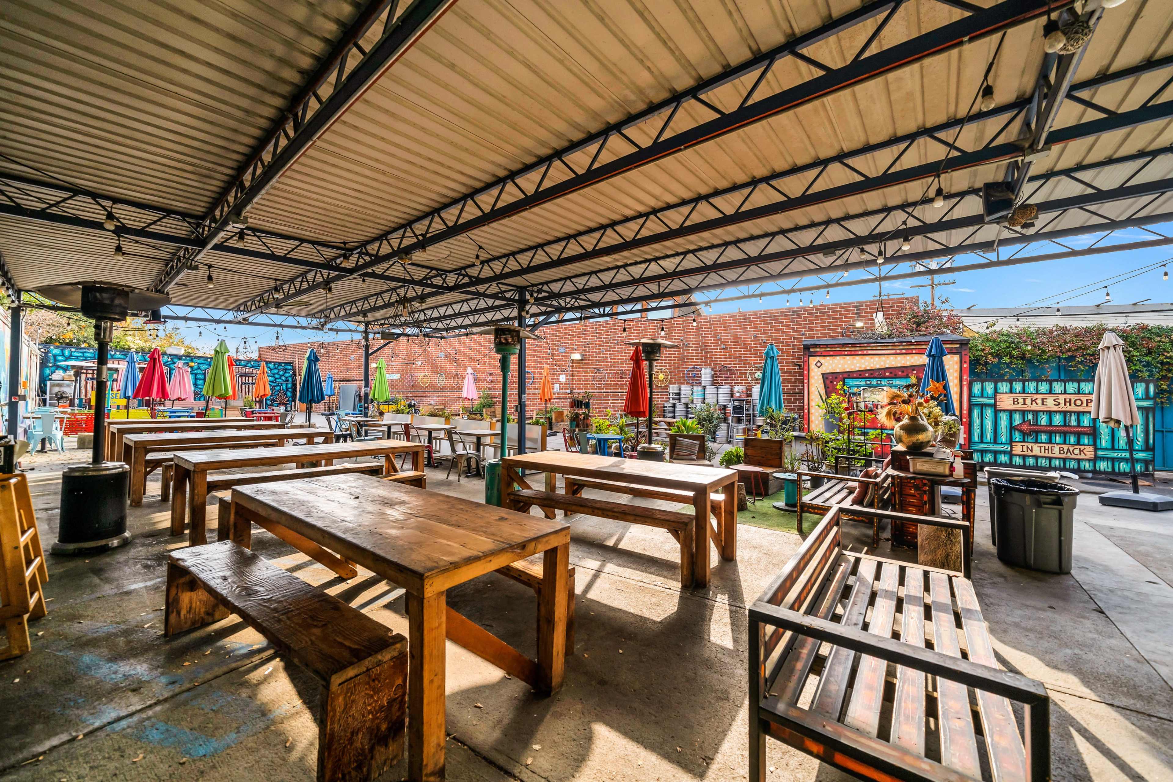 An outdoor seating area with wooden tables and benches beneath a metal roof, surrounded by colorful umbrellas and a brick wall in the background.