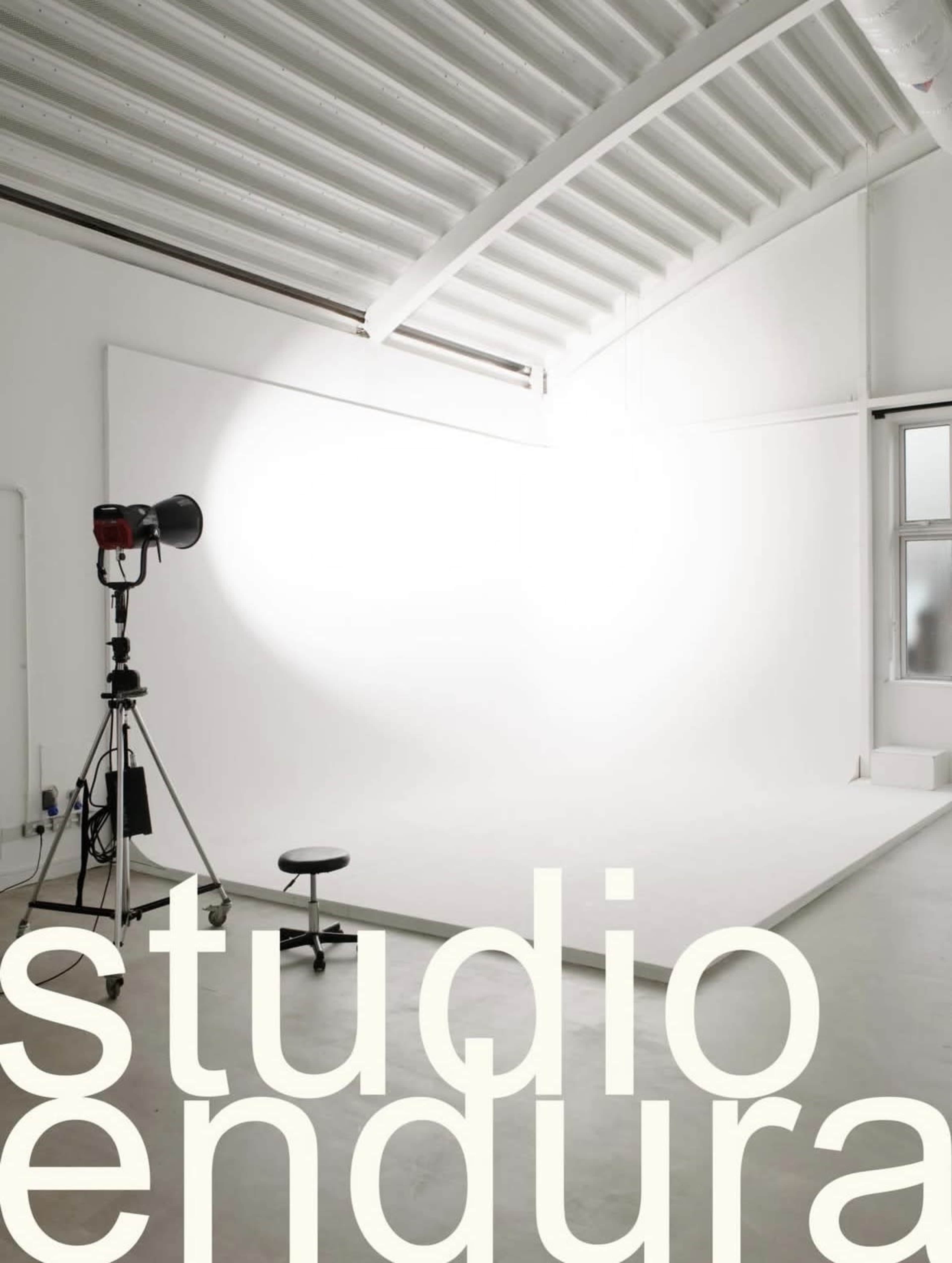 The image shows a minimalist photography studio with a white backdrop, a stool, and a spotlight on a stand.