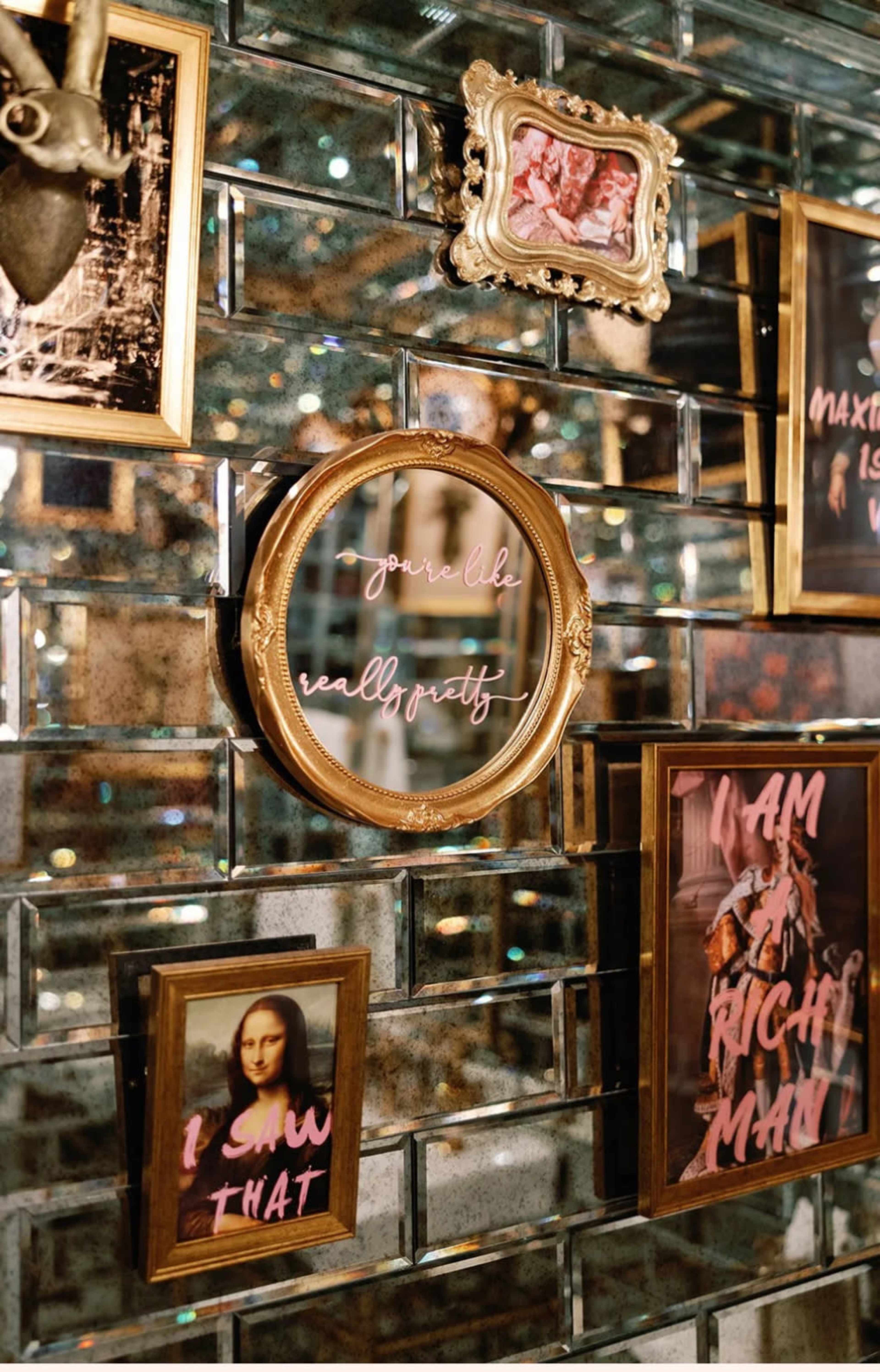 The image shows a wall adorned with various framed artworks and mirrors, including a circular mirror with the phrase "you're like really pretty" written in cursive.