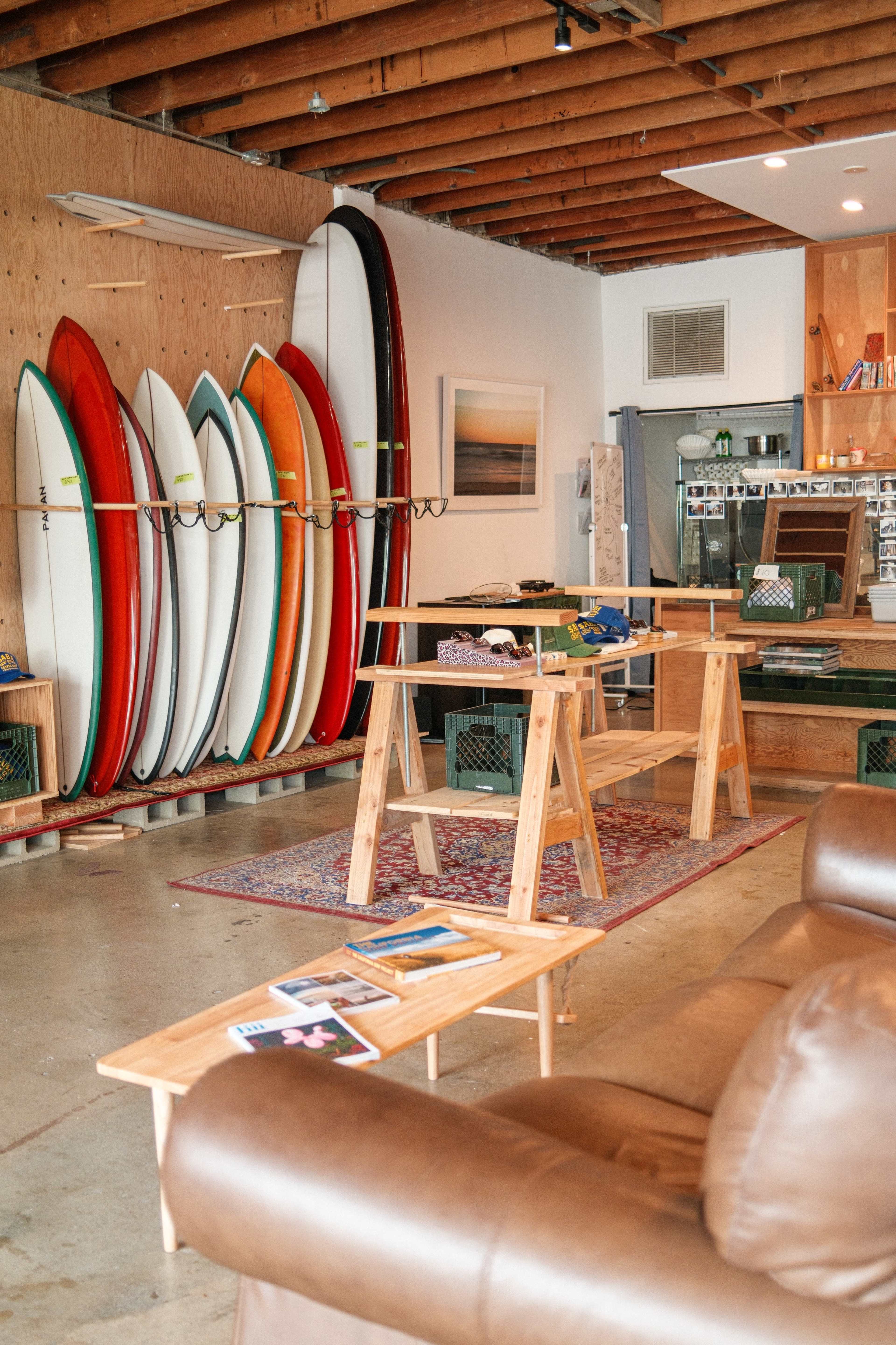 The image shows a surf shop interior featuring a row of colorful surfboards against a wooden wall, with wooden tables and a seating area in the foreground.