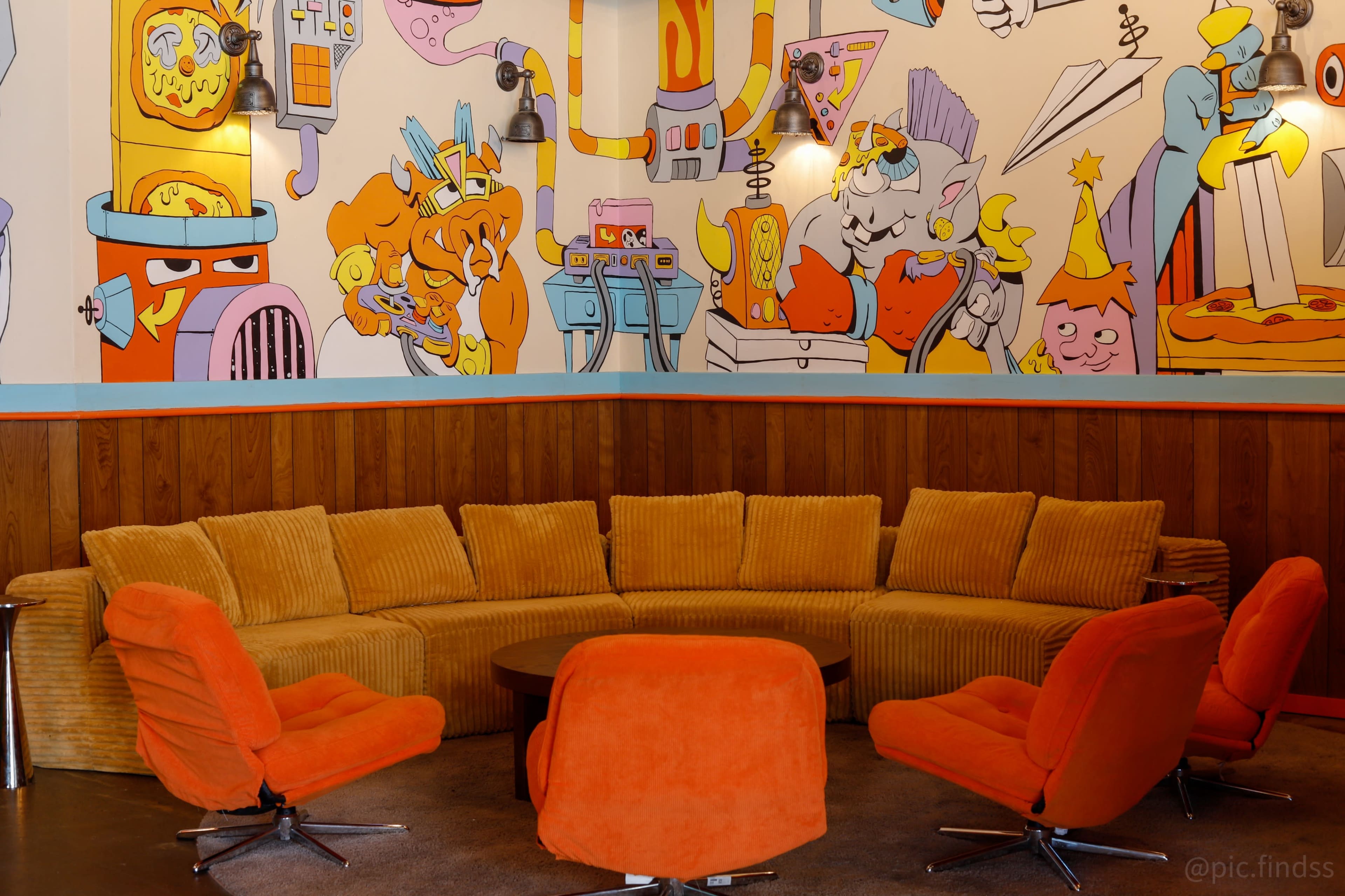 The image shows a circular seating arrangement made of orange upholstery in front of a vibrant, cartoon-themed mural on a wall.