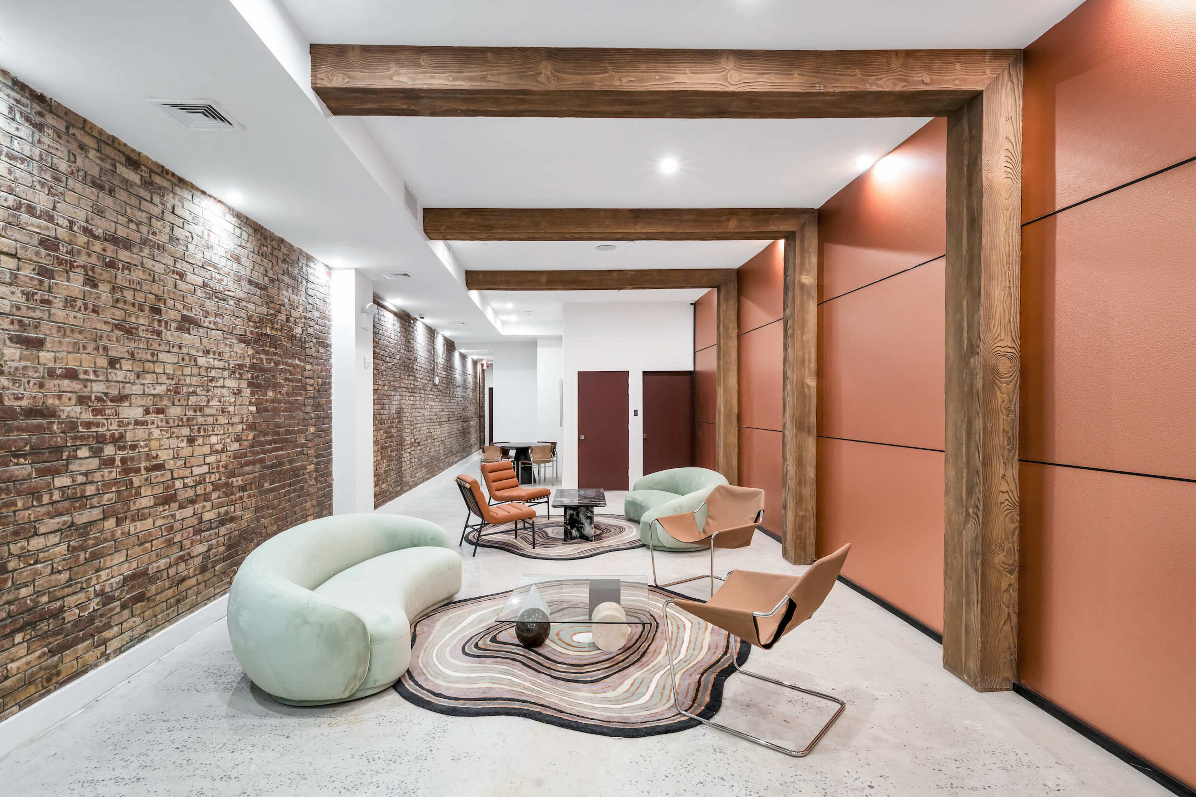 A modern interior corridor featuring a mix of exposed brick and orange paneling, with a seating area that includes a green sofa, brown chairs, and a coffee table atop a patterned rug.