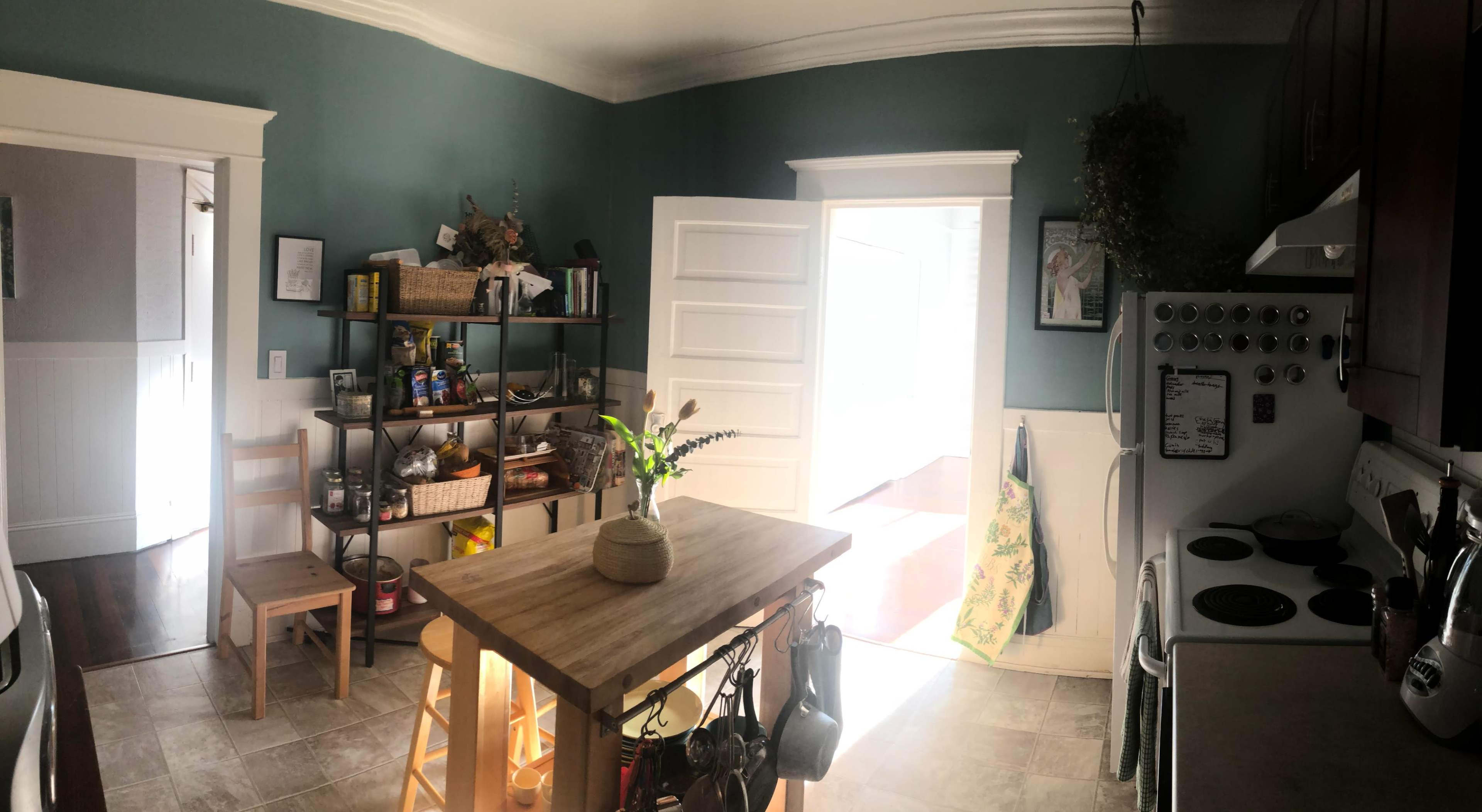 A kitchen with a wooden island, a shelf filled with jars and kitchen supplies, and an open door leading to a sunlit area.