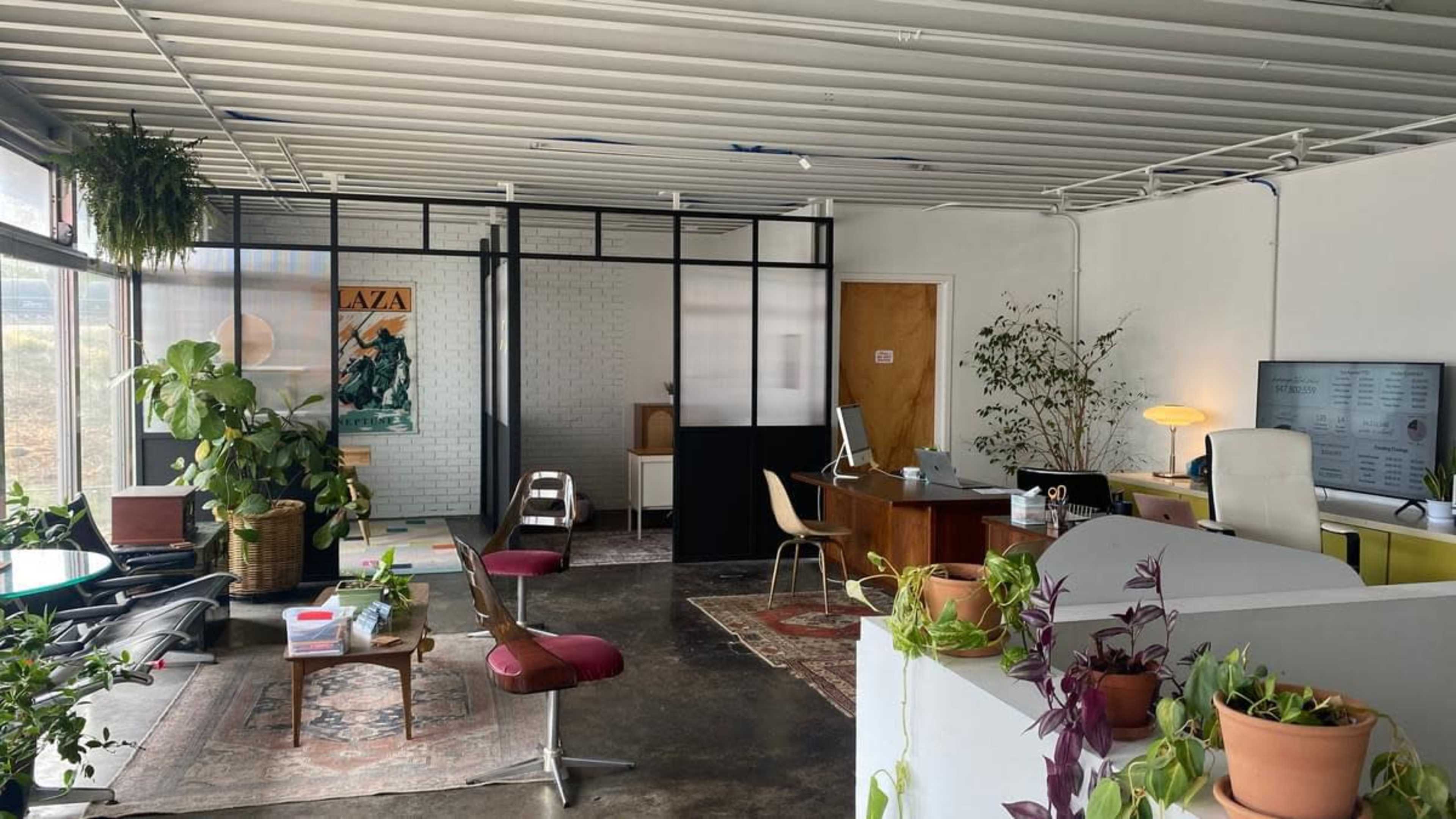 A modern office space with a combination of plants, furniture, and workstations, featuring a glass partition separating different areas.