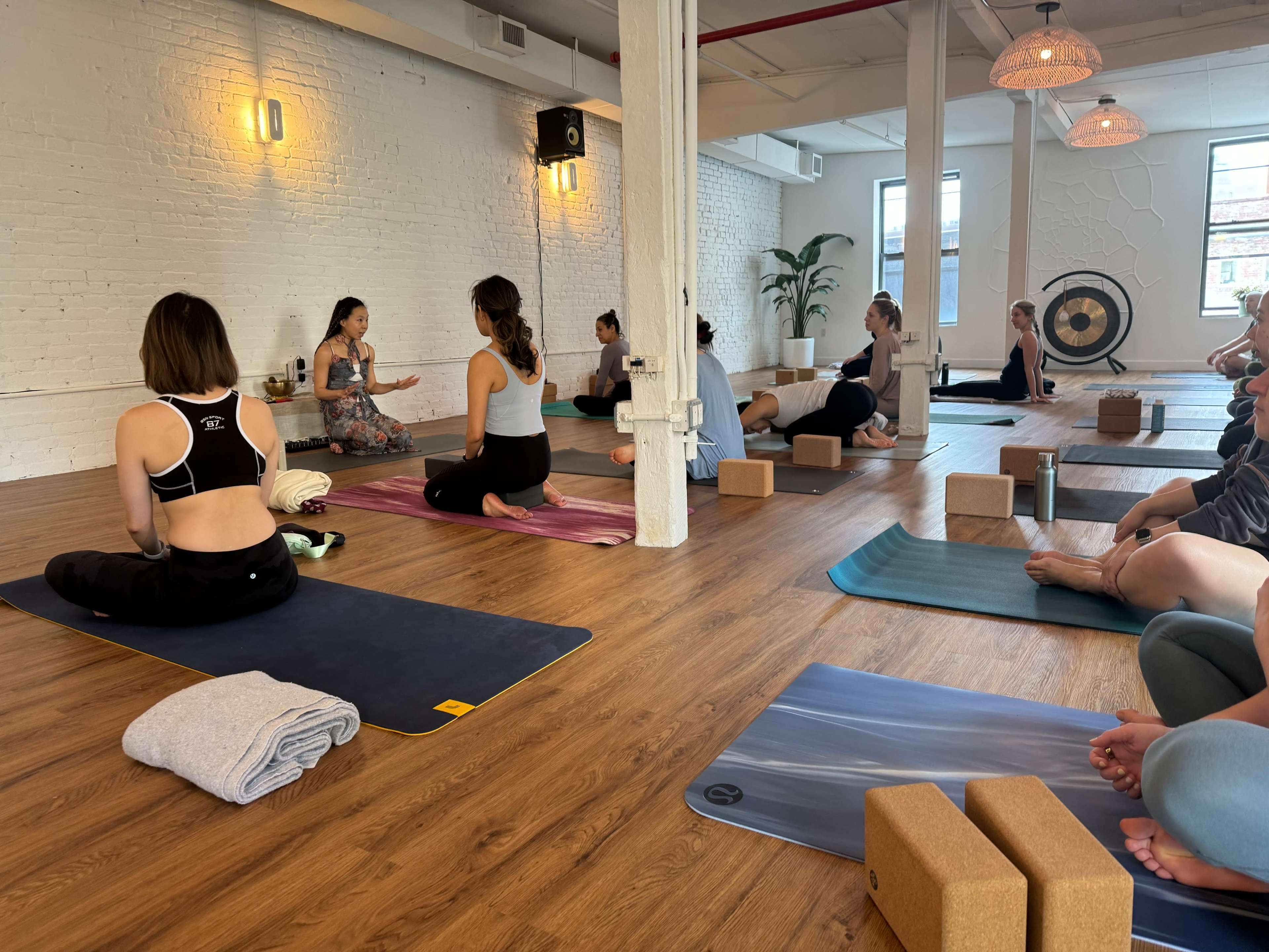 A group of individuals participates in a yoga class in a spacious studio with wooden flooring and large windows.
