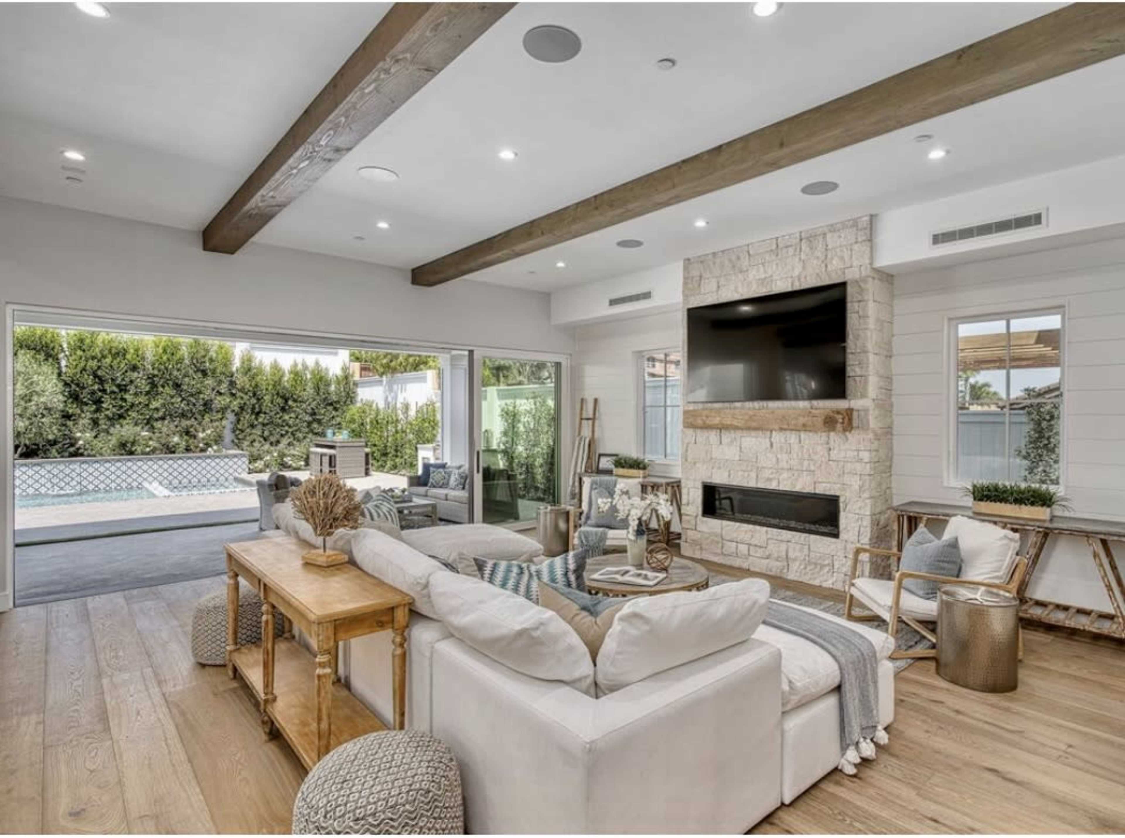 The living room features a light-colored sectional sofa, a stone fireplace, and large windows opening to an outdoor space with a pool.