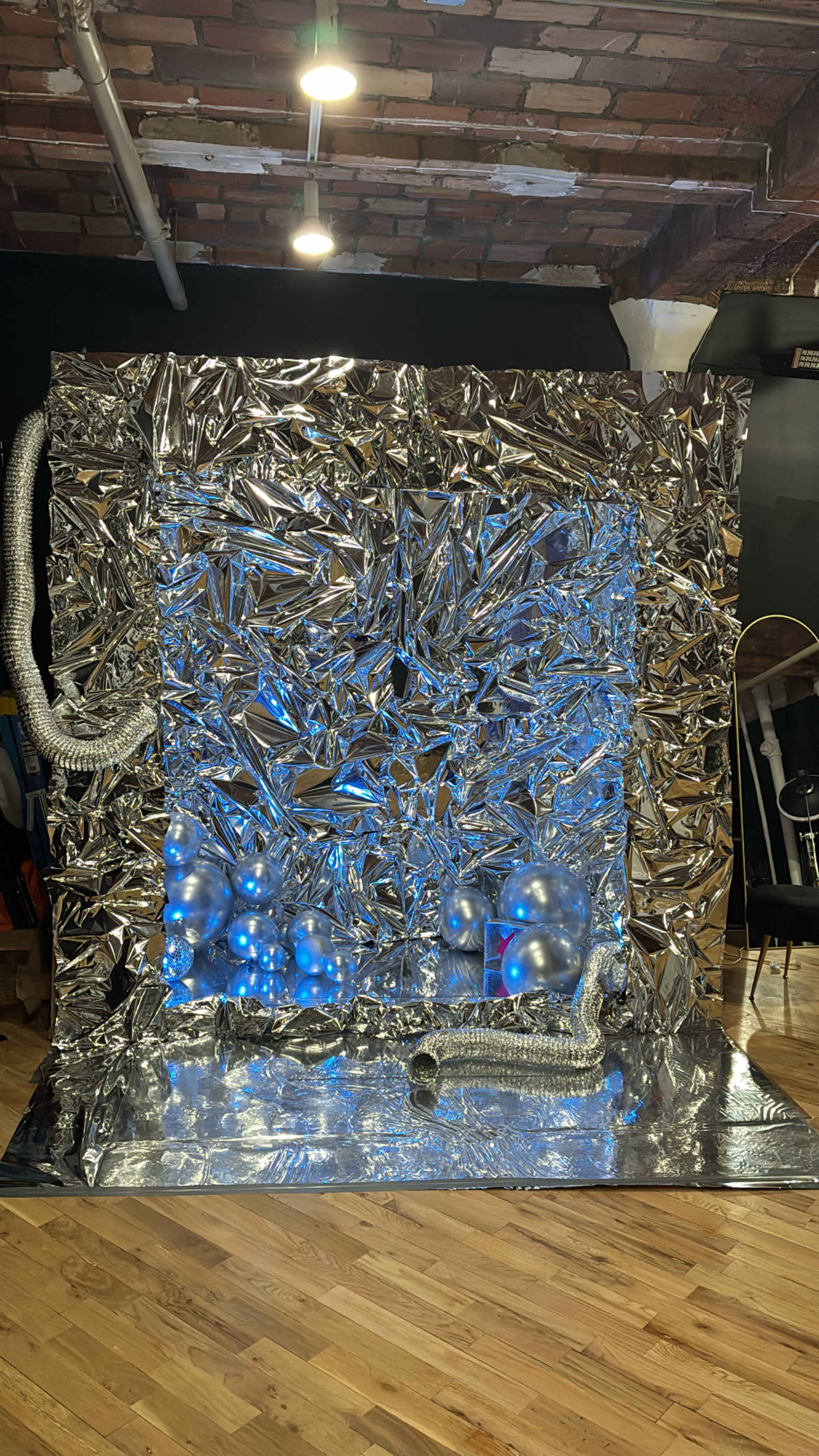 The image shows a shiny, crumpled metallic backdrop featuring blue lights and glossy balloons, with a textured surface creating a futuristic appearance.