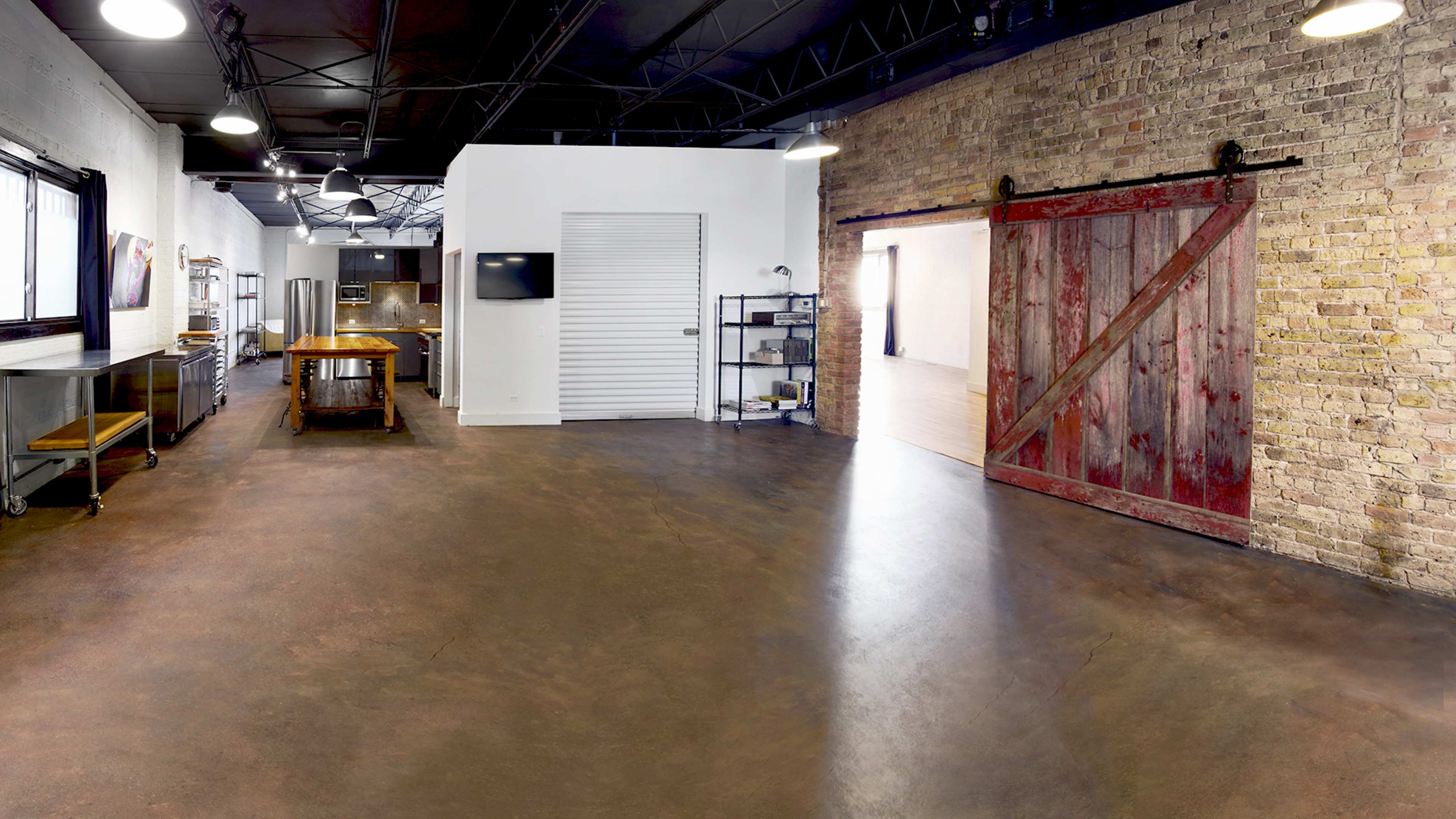 A spacious, modern kitchen area featuring a large wooden table, stainless steel appliances, and a rustic red barn door set against a wall of exposed brick.