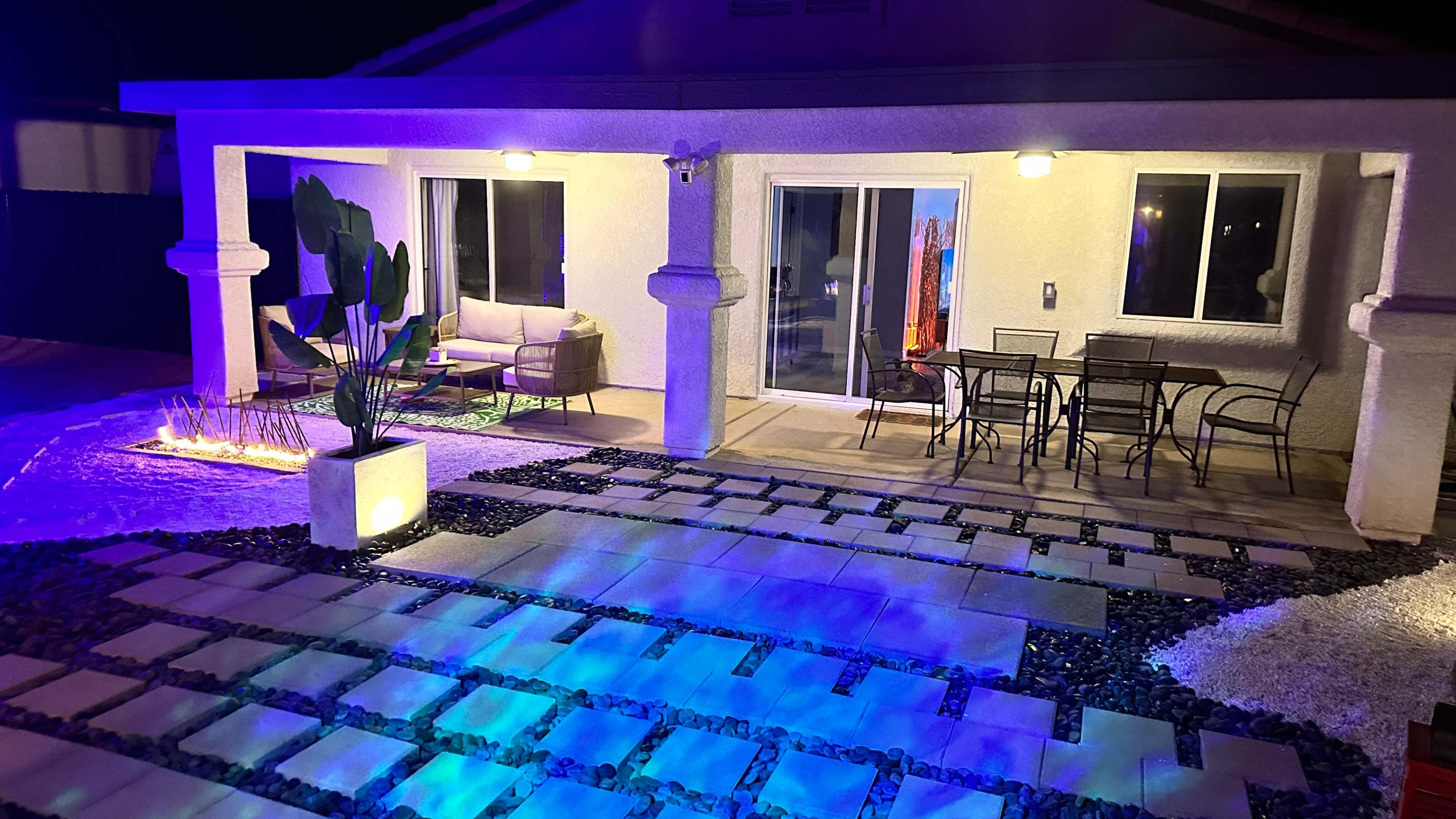 A well-lit patio area of a house with a seating arrangement and a dining table, surrounded by decorative landscaping.