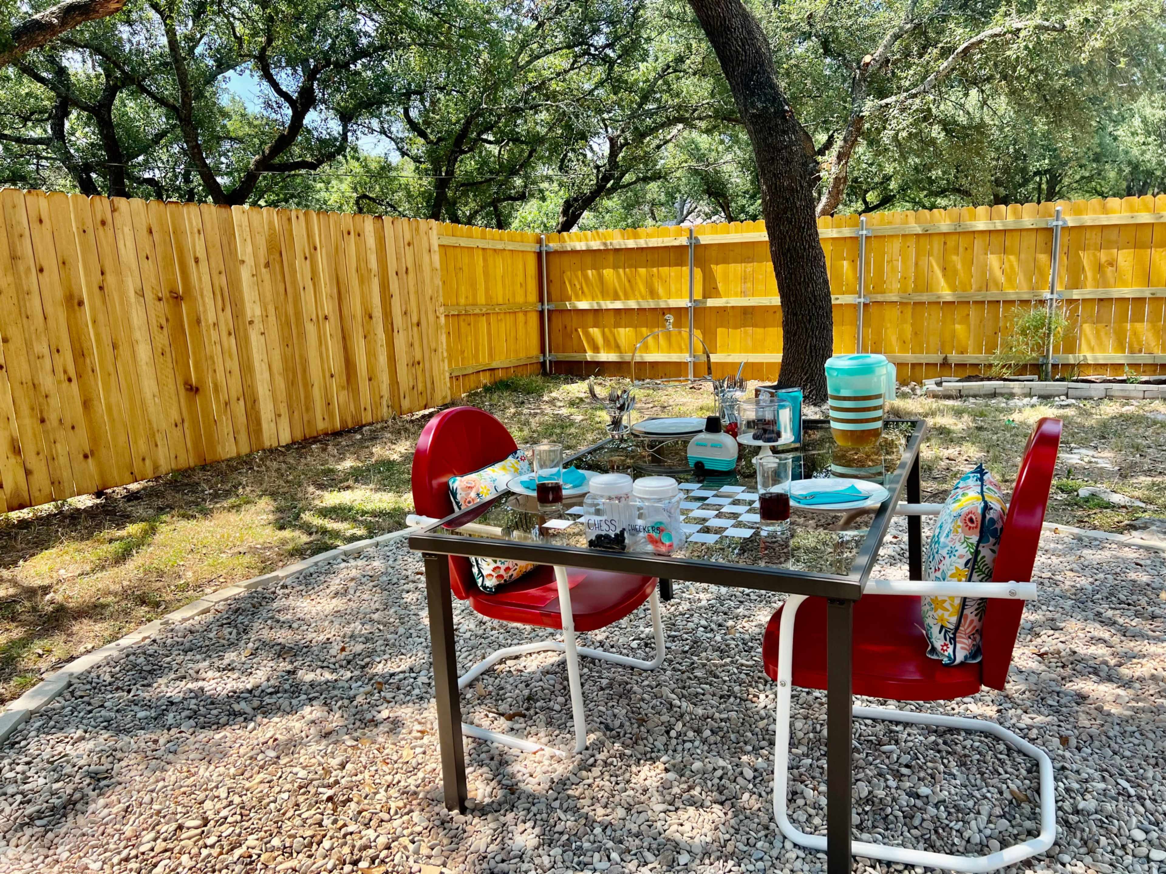 A table set with dinnerware and drinks is surrounded by two red chairs in a yard enclosed by a wooden fence.