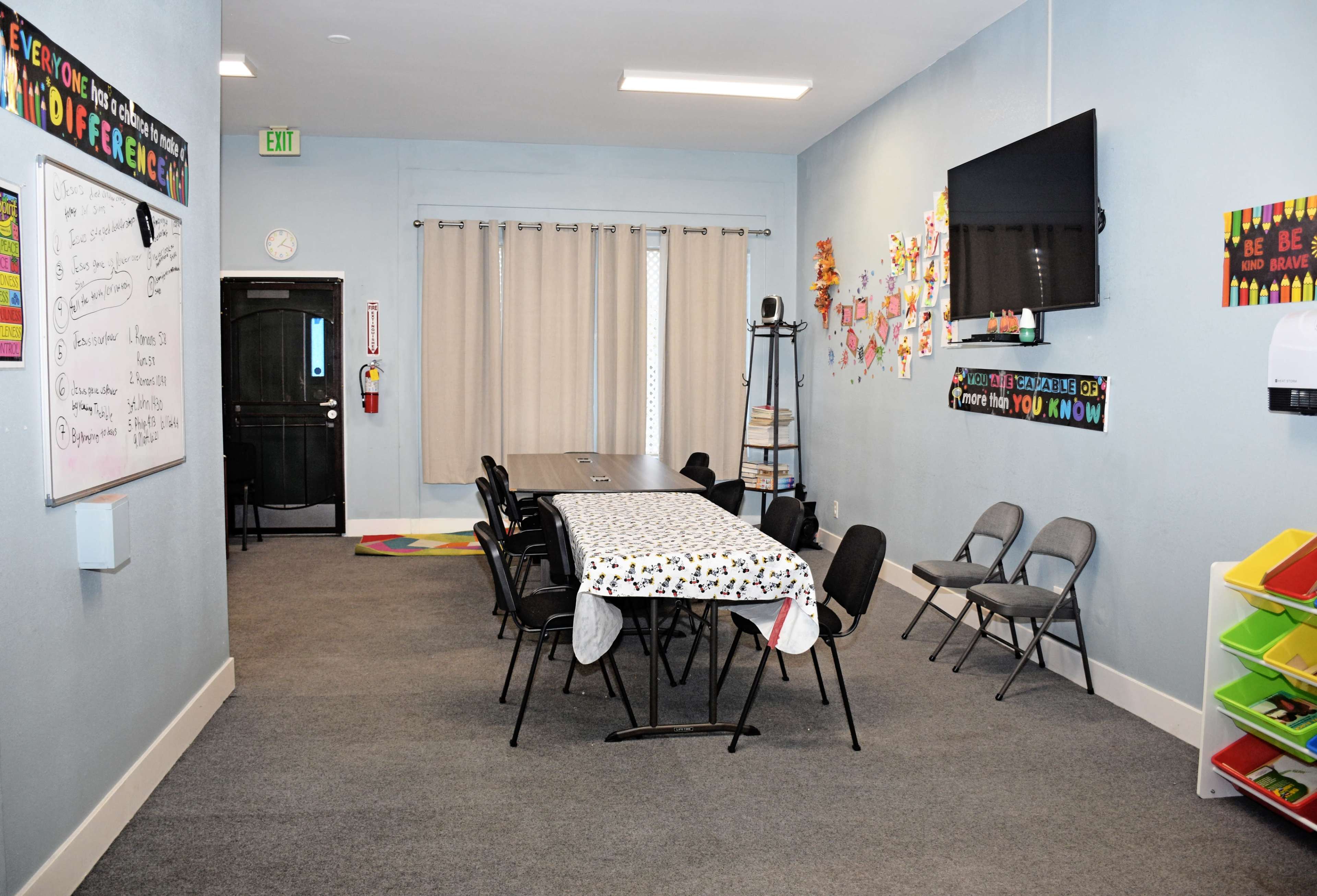 A classroom features a long table with chairs, a mounted television, and colorful educational decorations on the walls.