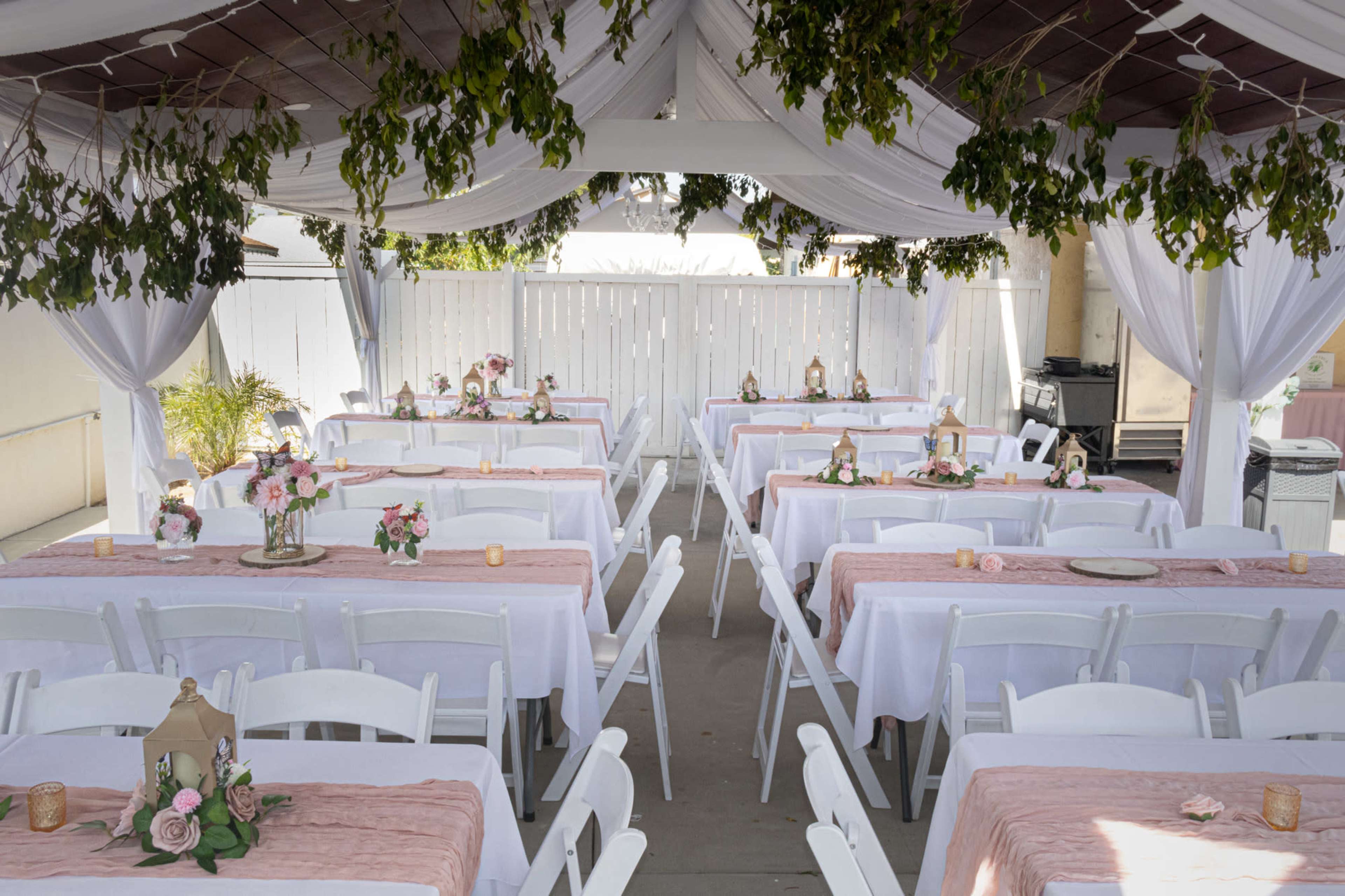 The image shows a series of long tables covered with white tablecloths and pink runners, arranged under a tent adorned with draped fabric and greenery.