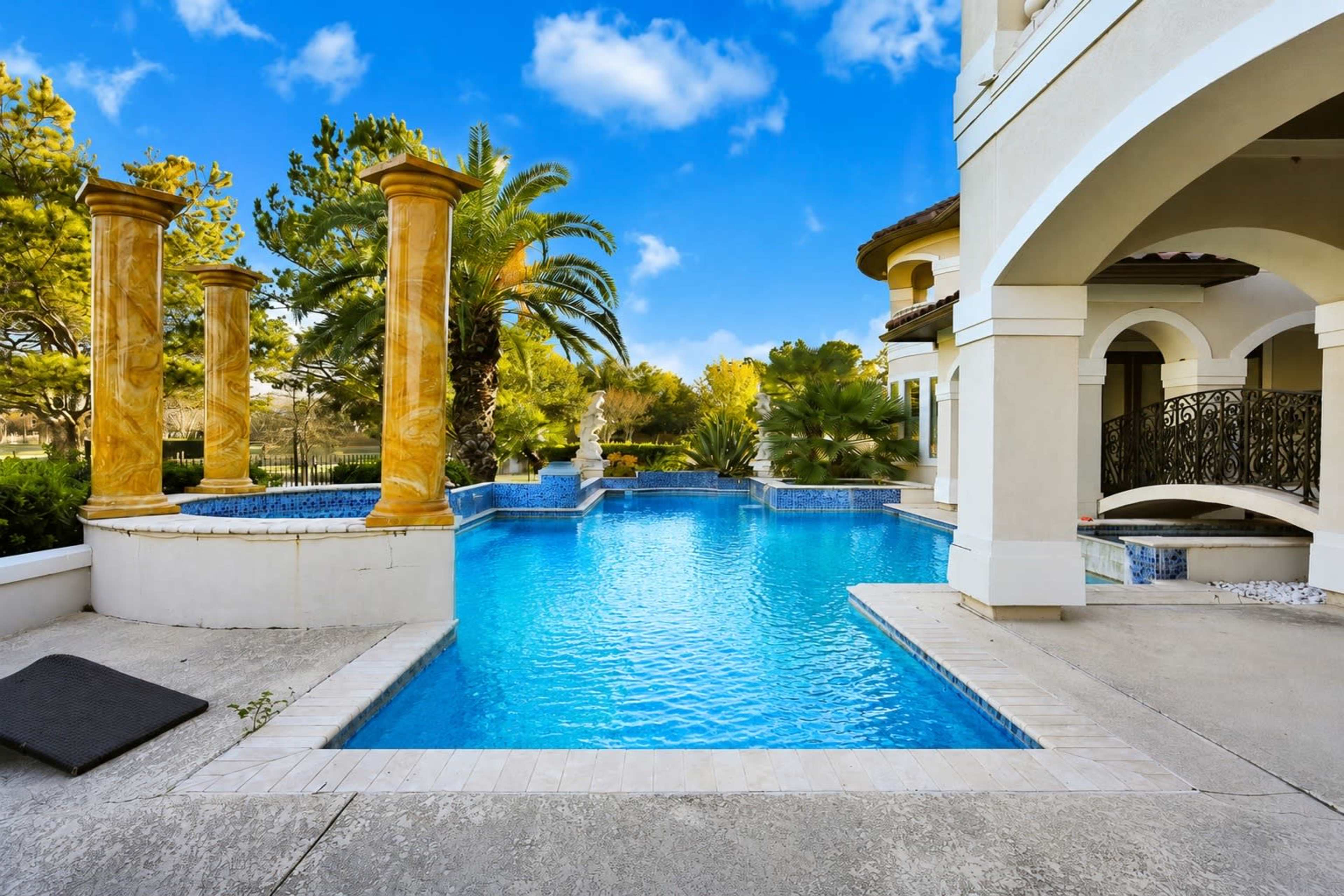 The image shows a luxurious swimming pool surrounded by palm trees and elegant columns, with clear blue water reflecting the sky.