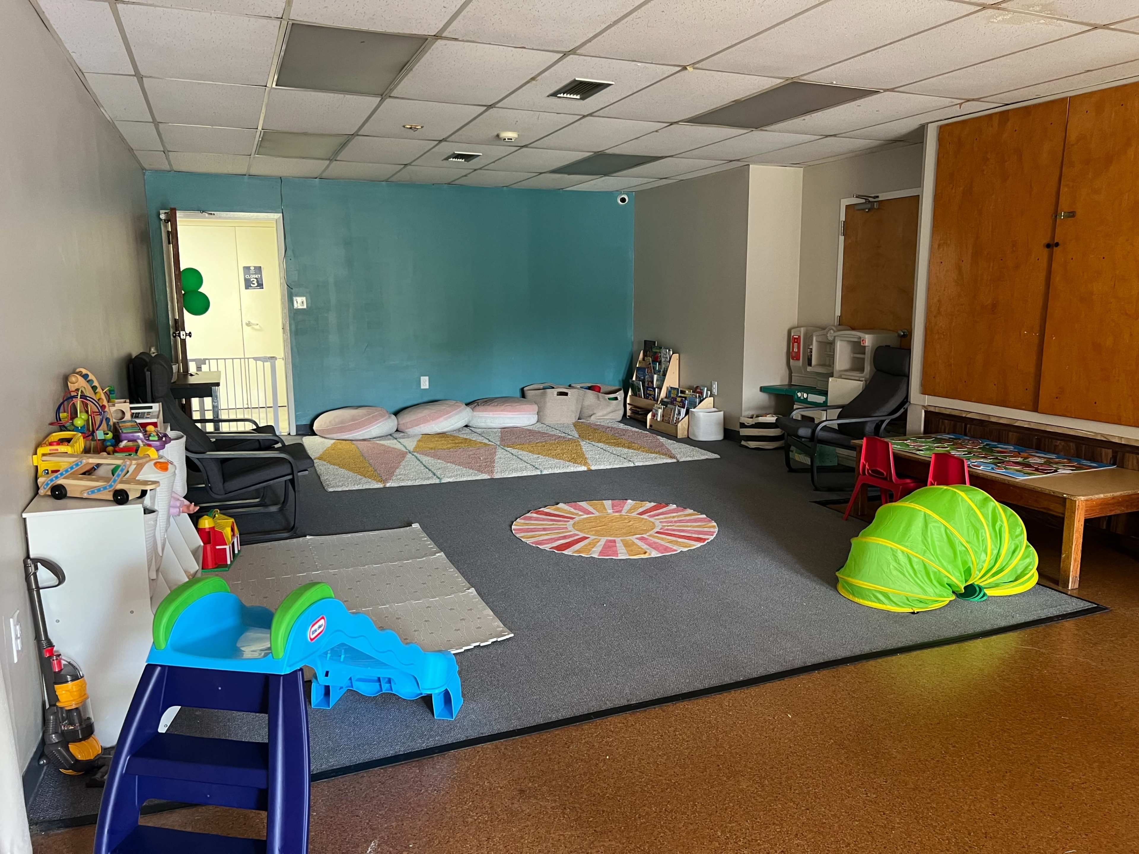 The image shows a spacious playroom with a colorful patterned rug, various children’s toys, two green tunnels, and a table with chairs in one corner.